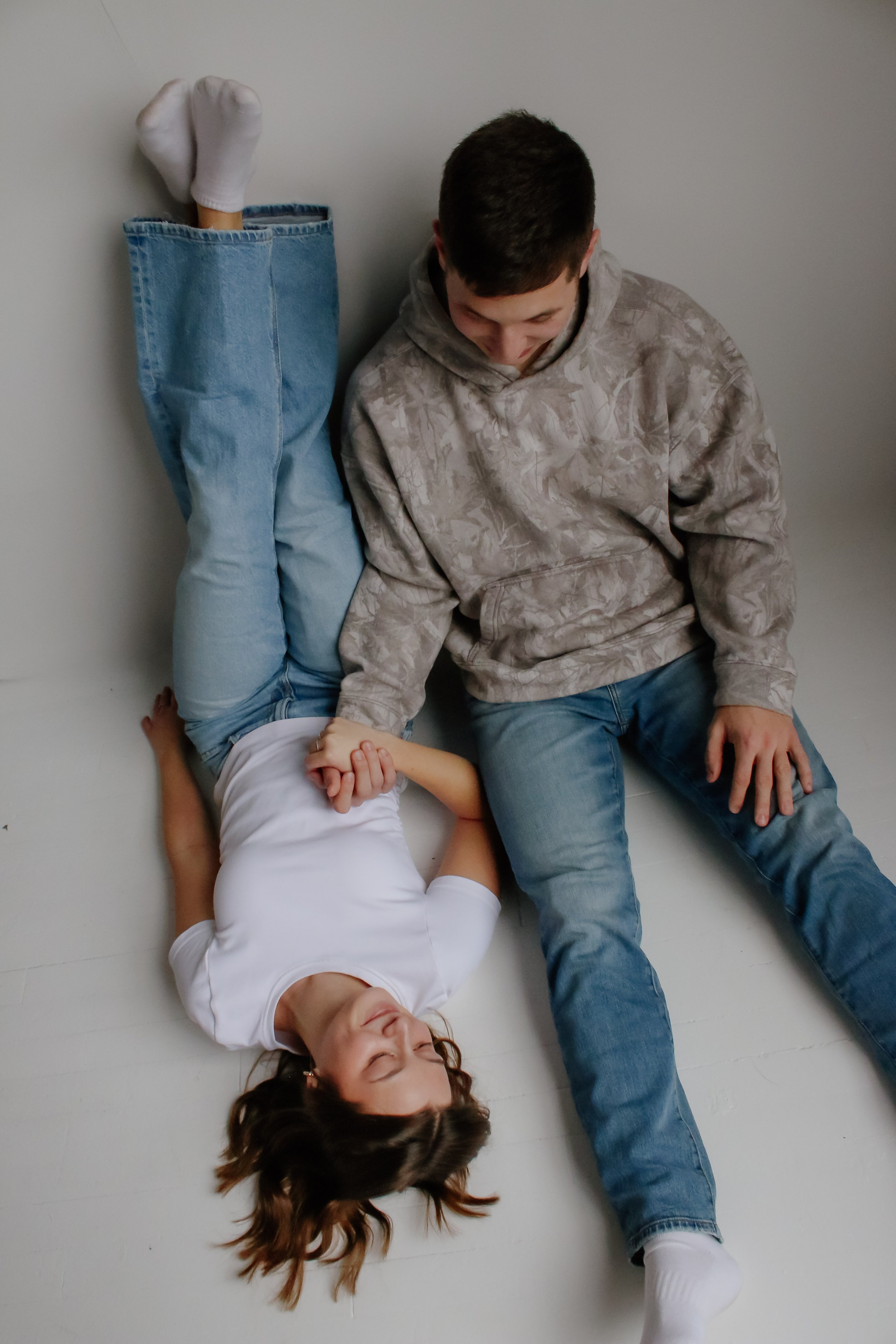 A young girl lying on the floor with her eyes closed, holding hands with an older boy sitting next to her, both smiling.