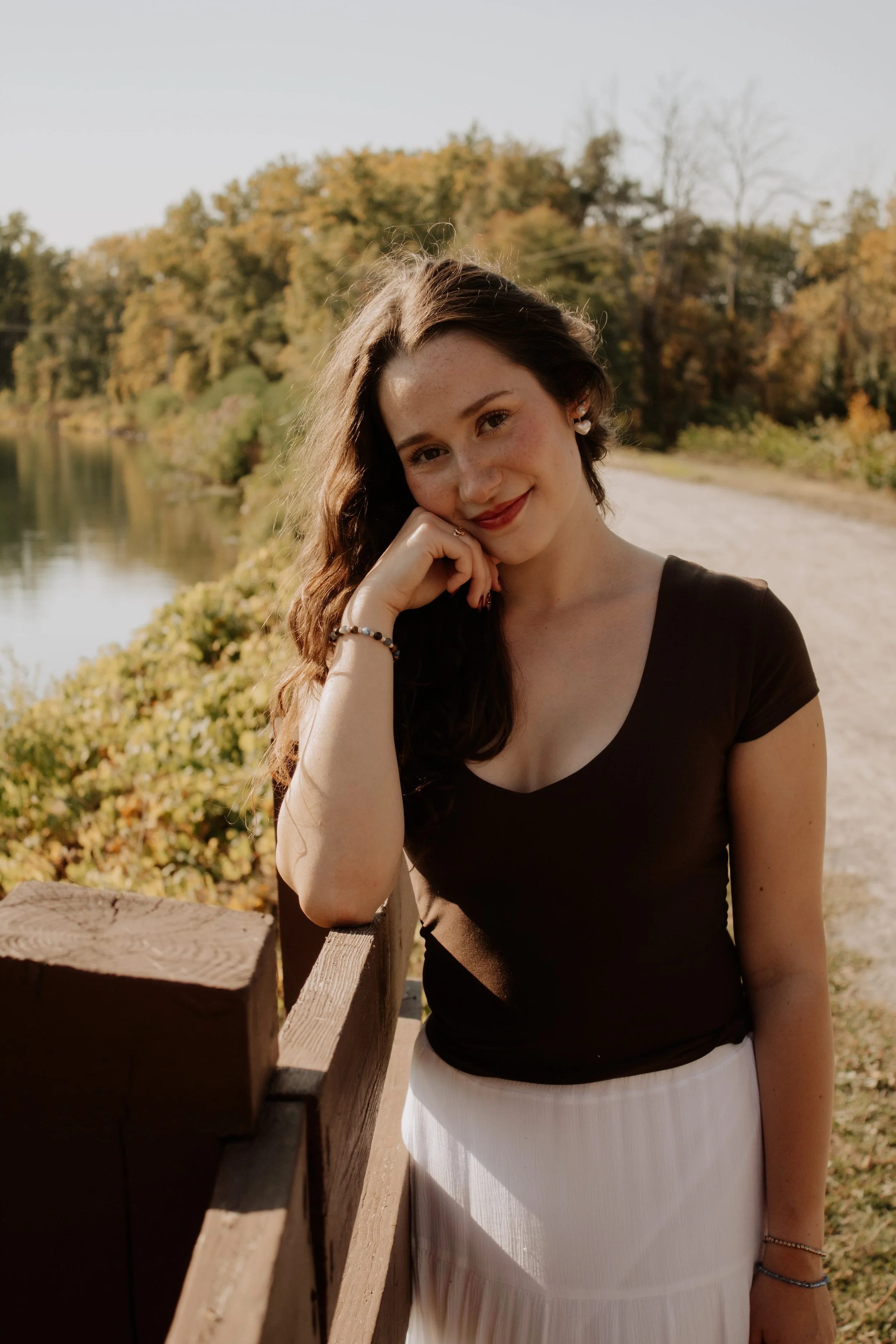 Young woman with dark hair and freckles leaning on a wooden railing along a lakeside path in autumn.