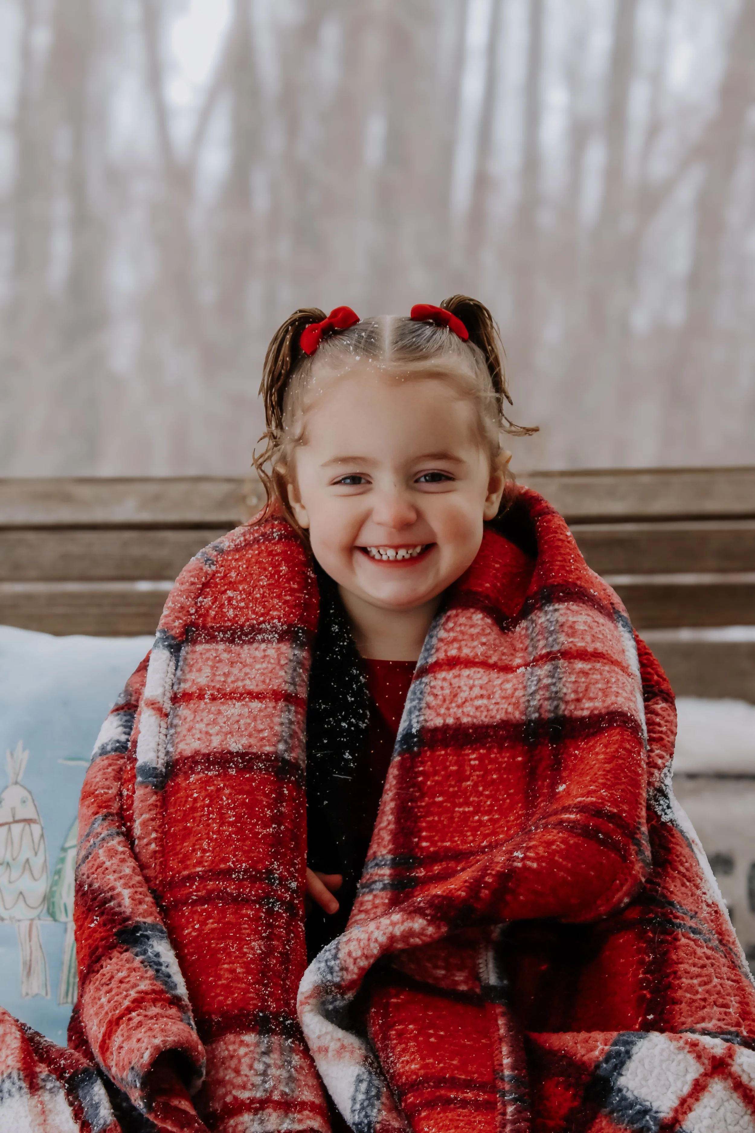 A smiling young girl with two red bows in her hair, wrapped in a red and black plaid blanket, sitting outdoors with a snowy background.