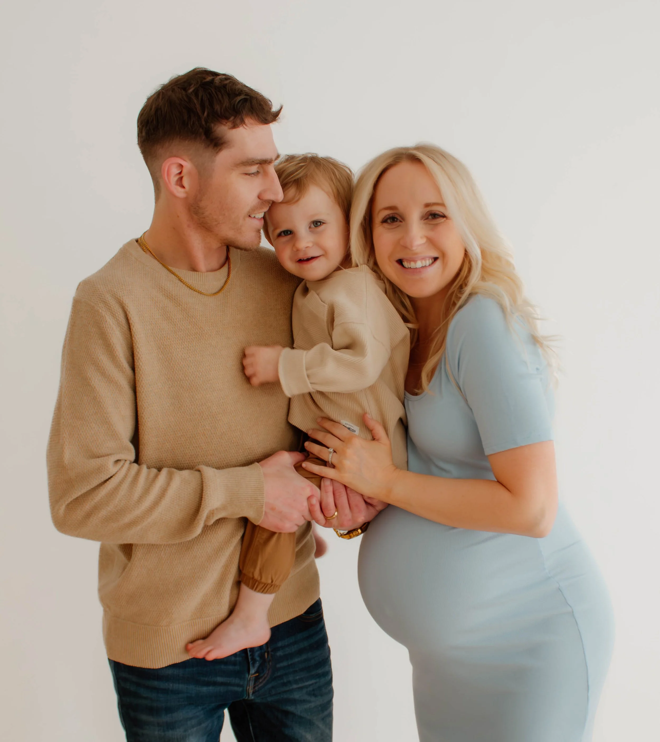 A smiling pregnant woman with blonde hair and a blue shirt, a man with short brown hair and a beige sweater, and a young child with light brown hair and a beige shirt, posing against a plain white background.