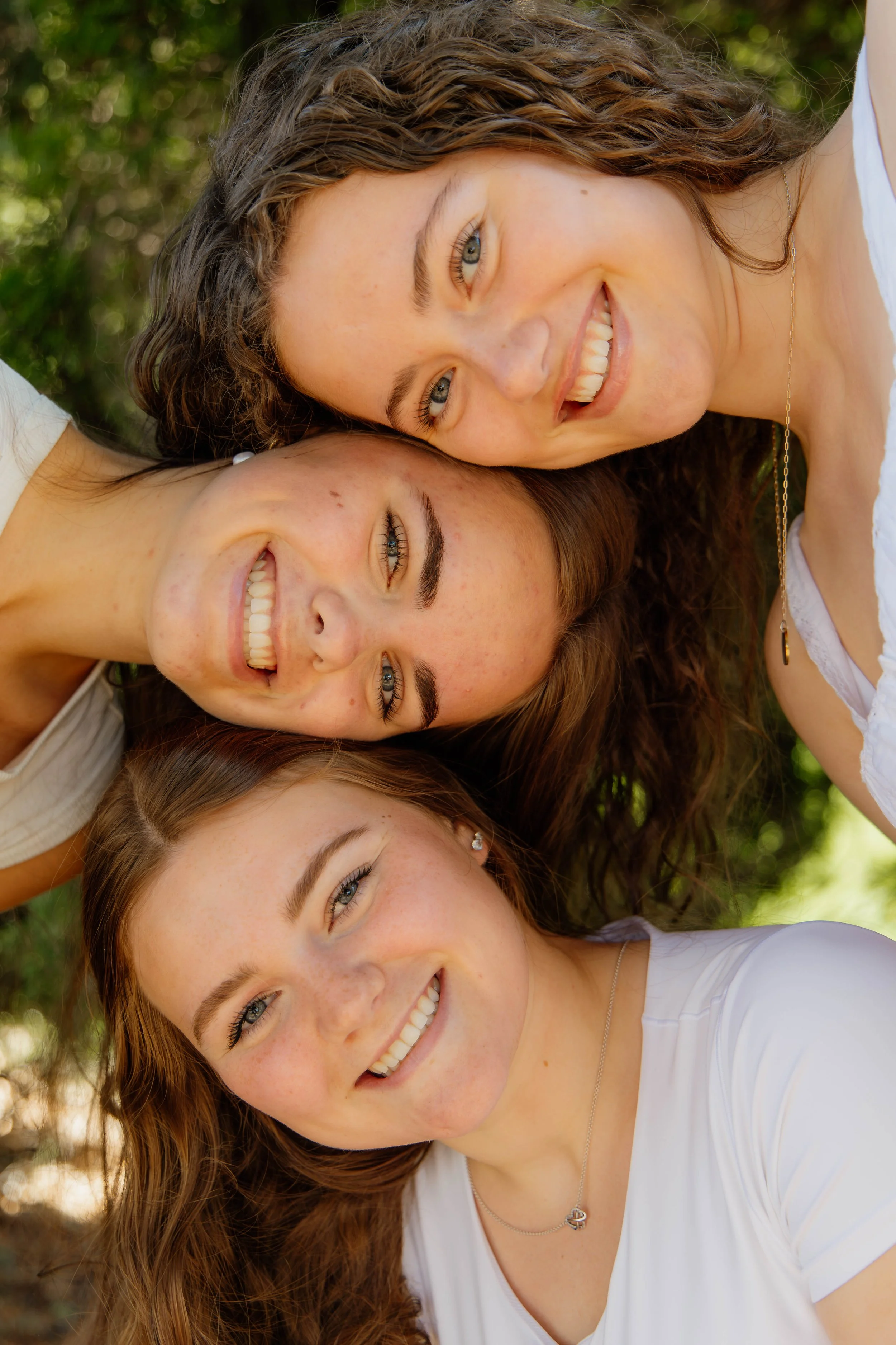 Close-up of four smiling women lying on the grass outdoors, with their heads together, in a sunny setting with blurred green foliage in the background.