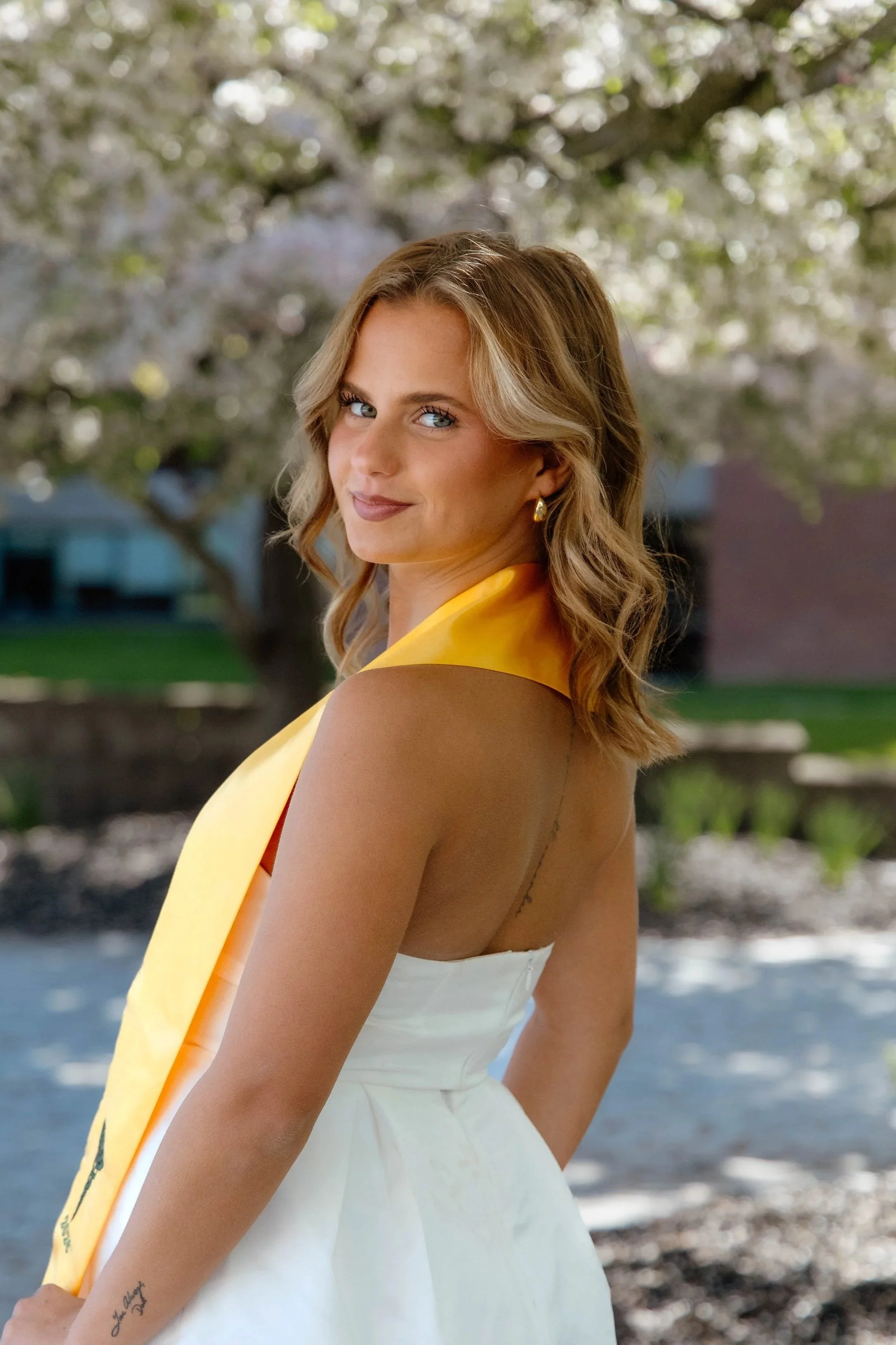 A young woman with wavy blonde hair, blue eyes, and light makeup, wearing gold earrings and a sleeveless white dress, stands outdoors in front of a blossoming tree with white flowers. She has a yellow scarf draped over her shoulder and is smiling softly at the camera.