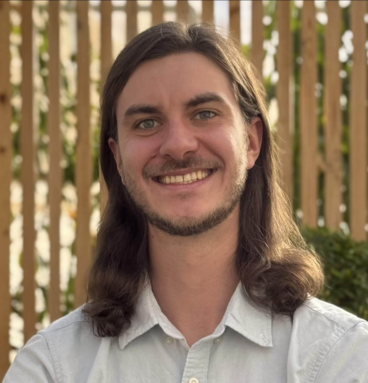 A man smiling outdoors with a wooden fence in the background.