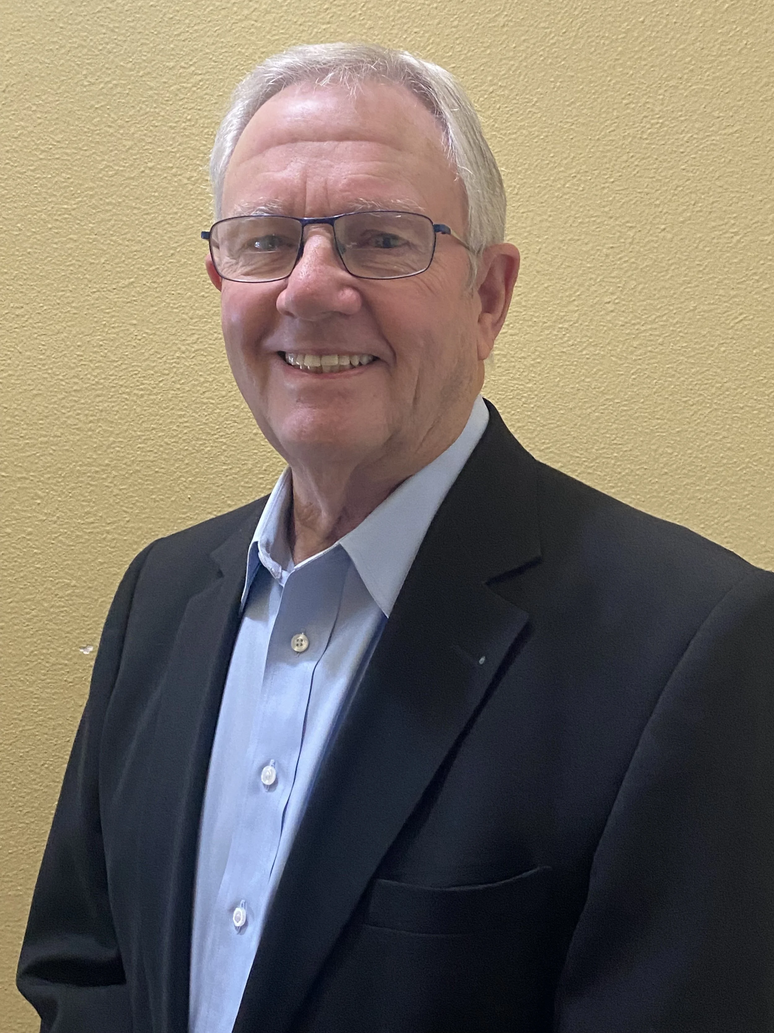An older man with gray hair and glasses, smiling, wearing a black suit jacket and a light blue dress shirt, standing against a yellow textured wall.