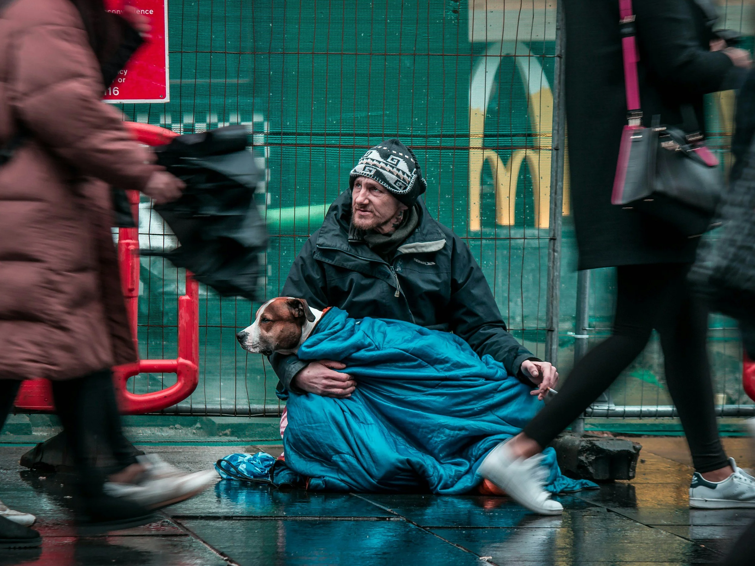 A man sitting on the wet ground with a dog wrapped in a blue blanket, surrounded by blurred pedestrians in an urban setting with a green fence and McDonald's sign in the background.