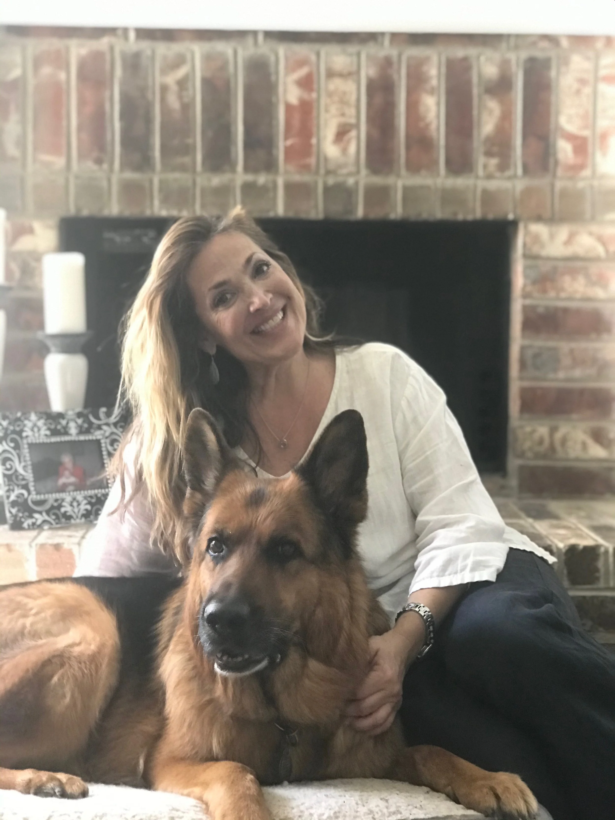 A woman smiling and leaning close to a large German Shepherd dog lying on a rug in front of a brick fireplace with a framed photo and candles on the mantle.