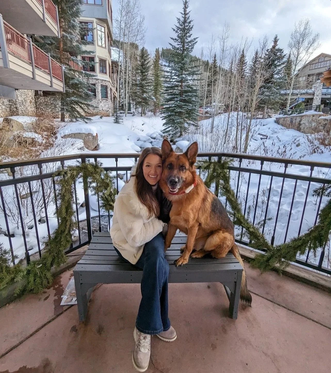 A woman in a white jacket sitting on a bench with a large German Shepherd dog in a snowy outdoor setting, with trees and buildings in the background.