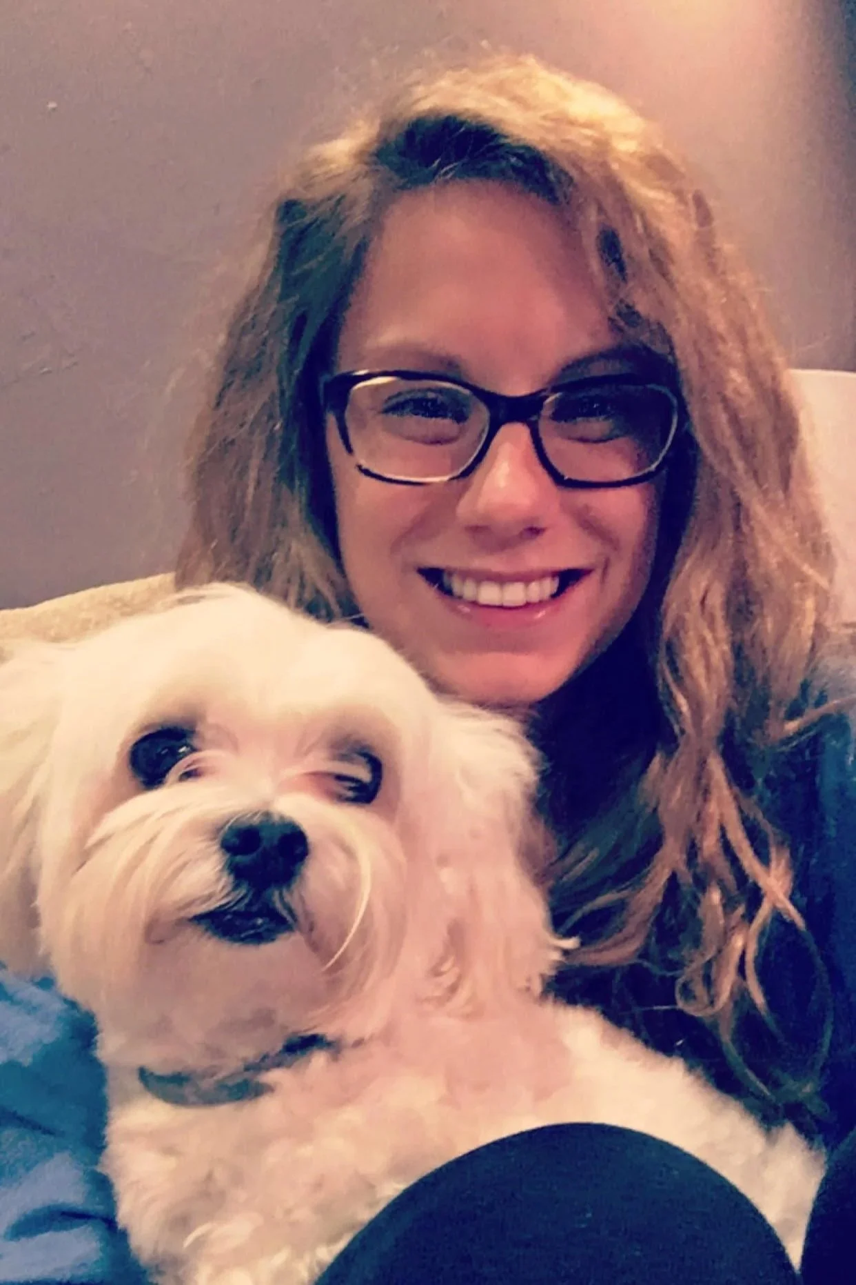 A woman with glasses and wavy hair smiling with a small white dog with dark eyes and a black nose.