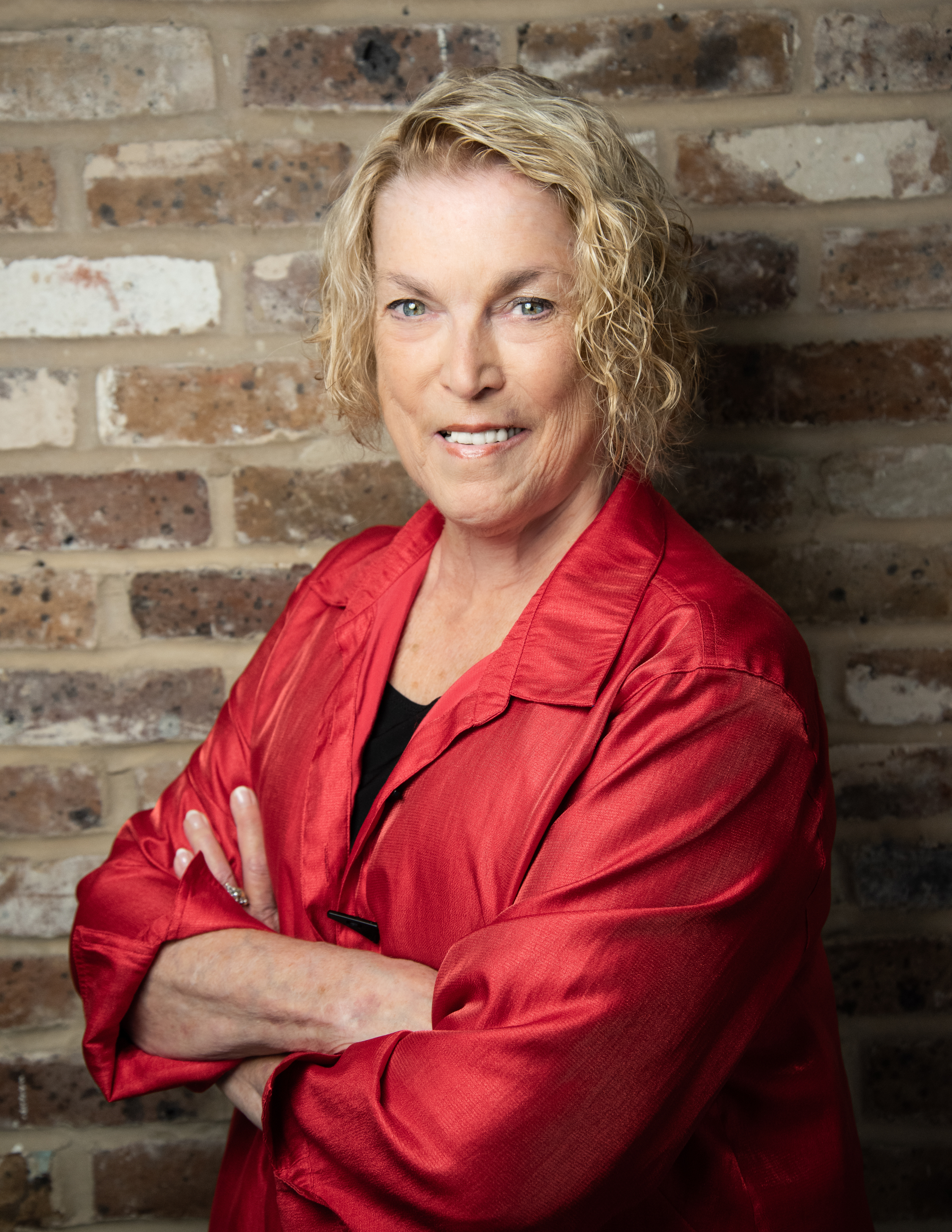 A woman with curly blonde hair wearing a red satin blouse, standing with her arms crossed against a brick wall, smiling at the camera.