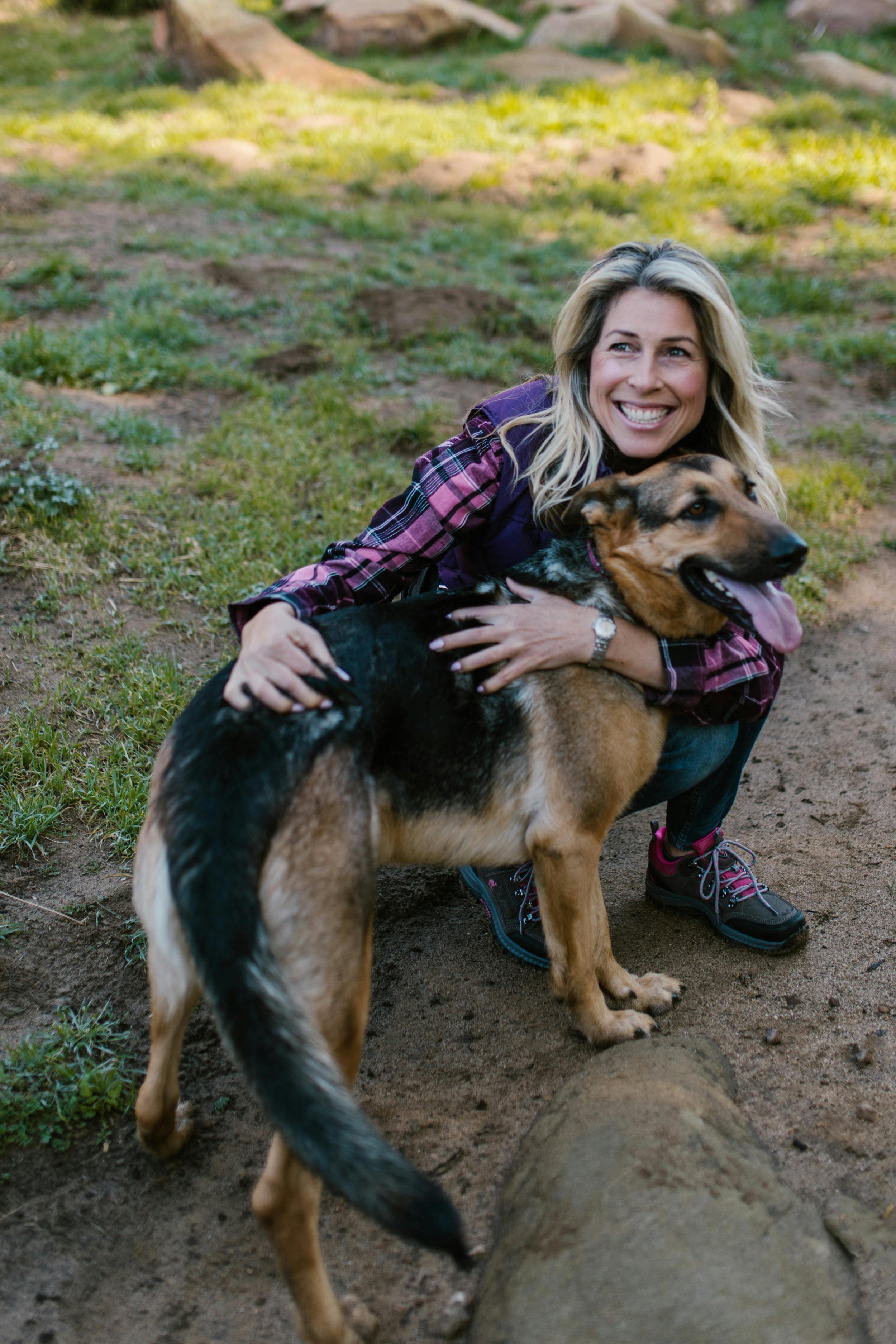 A woman with blonde hair smiling and hugging a large service dog outdoors on a patch of dirt and grass.