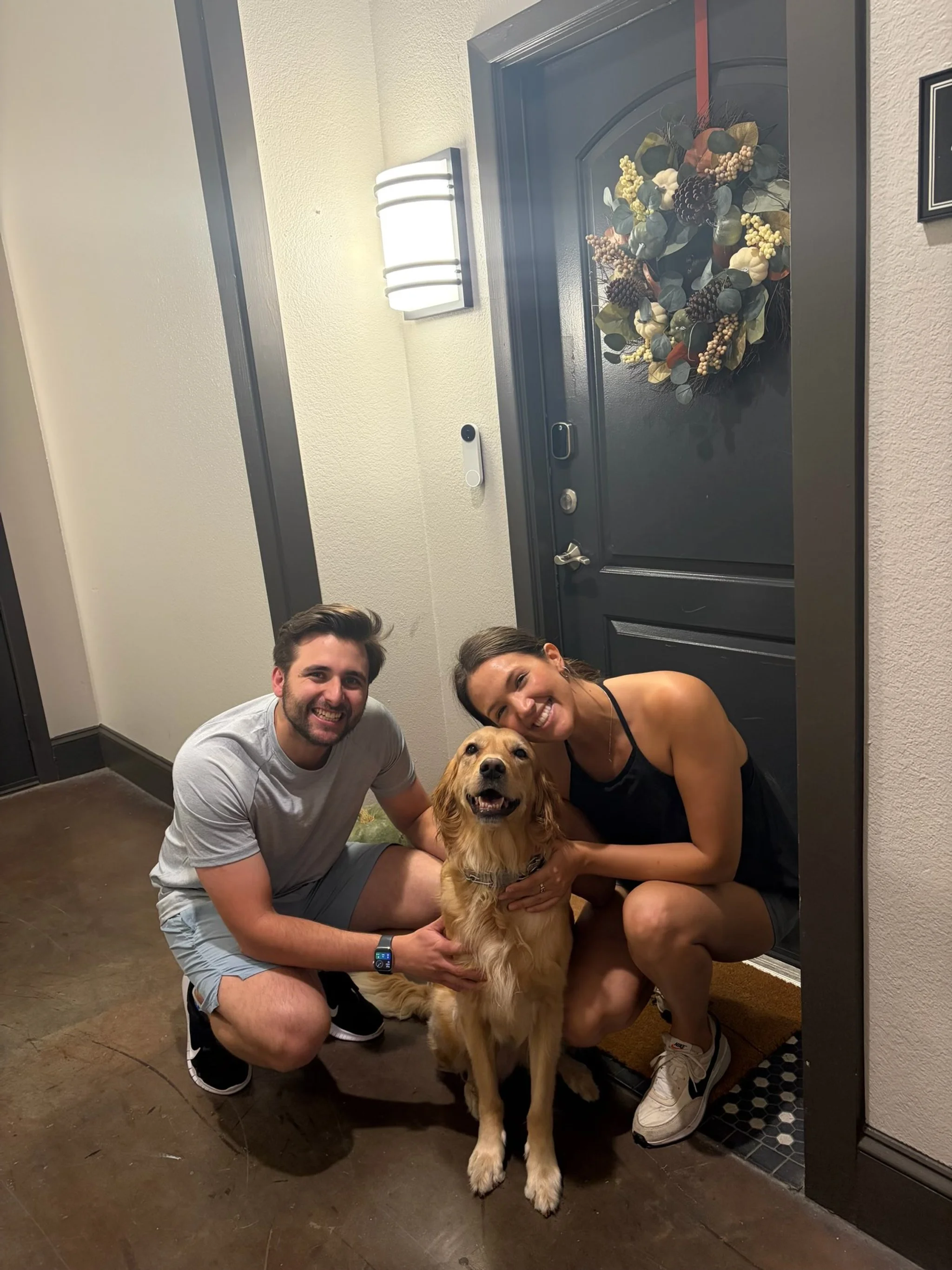 A smiling man and woman squatting down with a golden retriever dog in front of a dark entry door decorated with a fall-themed wreath inside a home. The woman and man are holding the dog, and the woman is hugging the dog.
