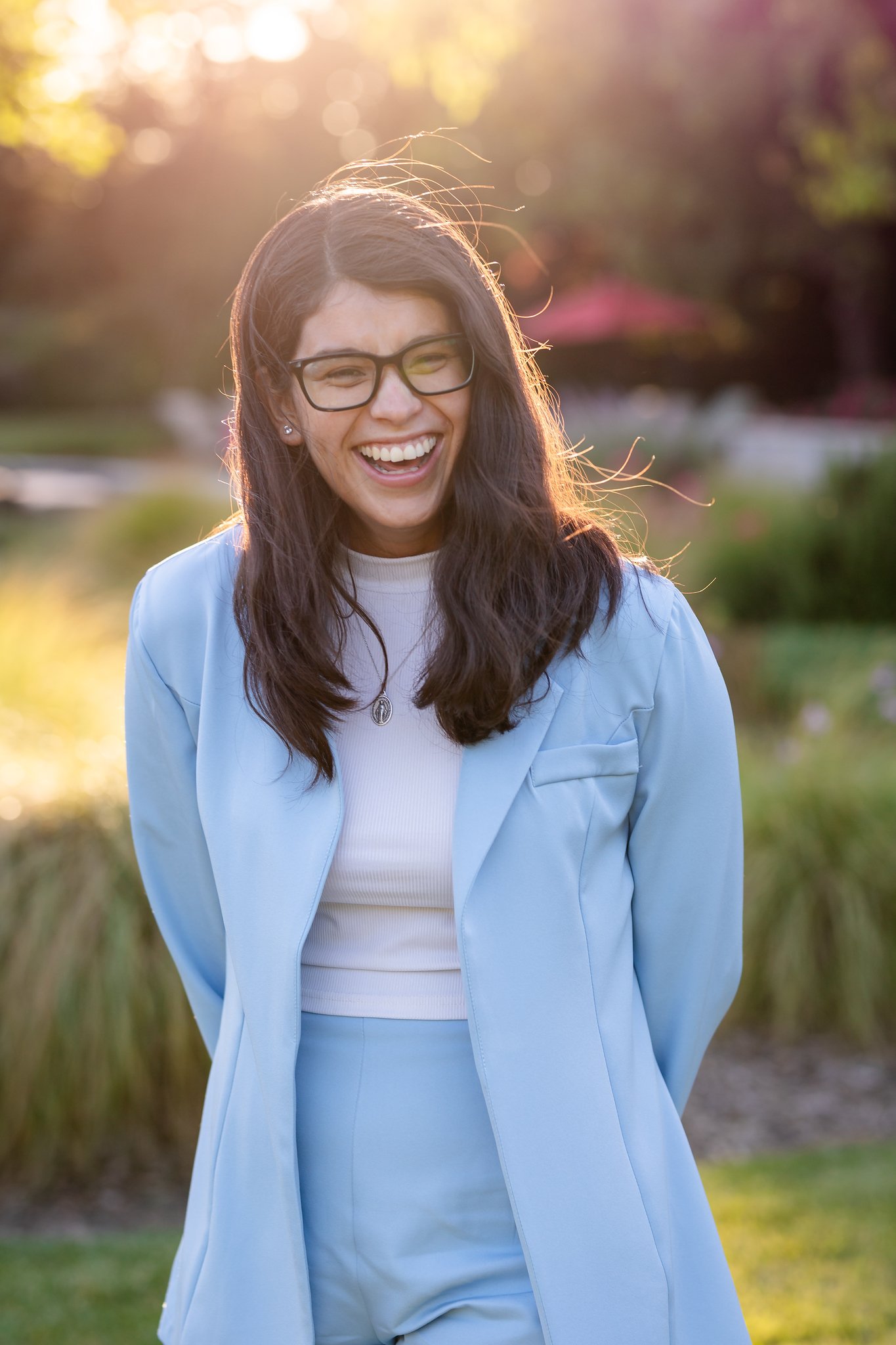 Woman with glasses smiling outdoors in sunlight, wearing a light blue blazer and pants, with a necklace and earrings, in a garden setting.