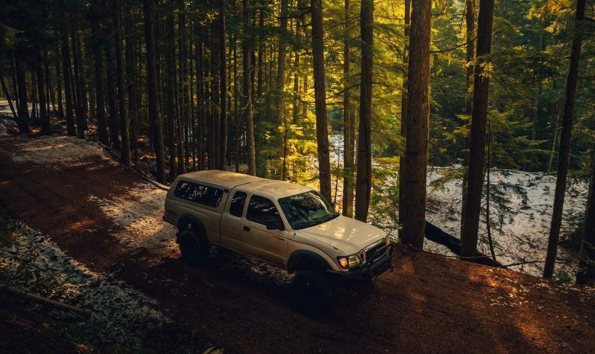 A white pickup truck with a camper shell driving on a dirt trail through a dense forest with tall trees and a river visible through the trees.