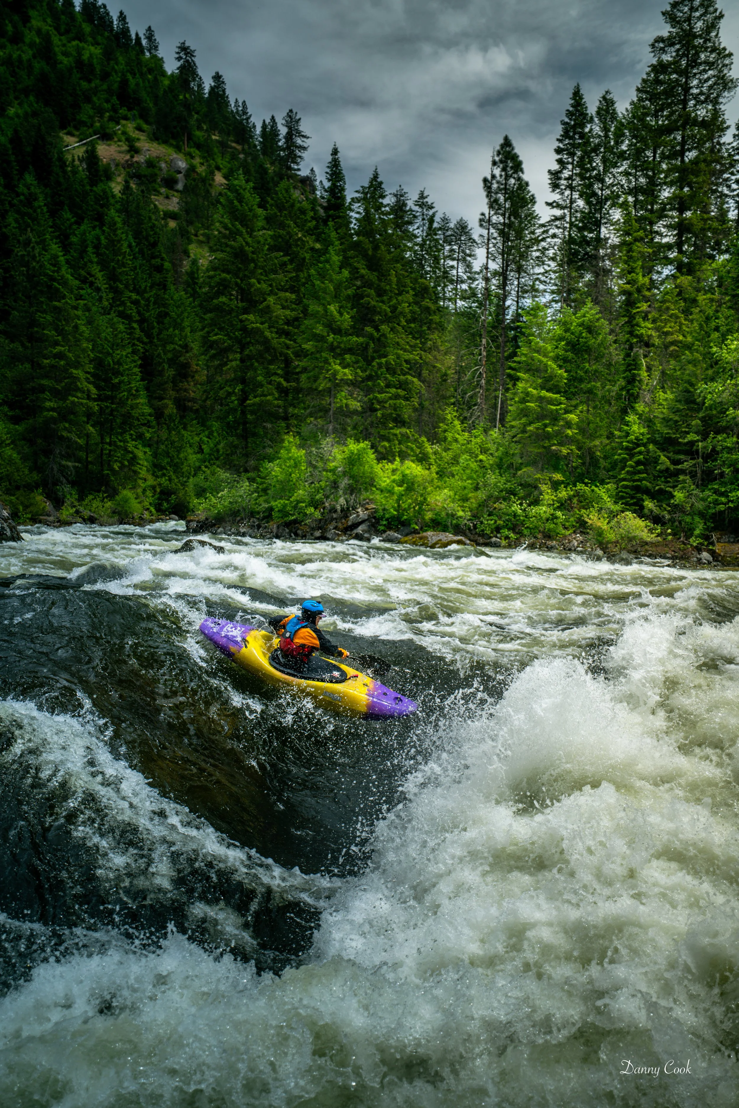 A person kayaking on a fast-moving river surrounded by lush green trees and a forested mountainside under a cloudy sky.