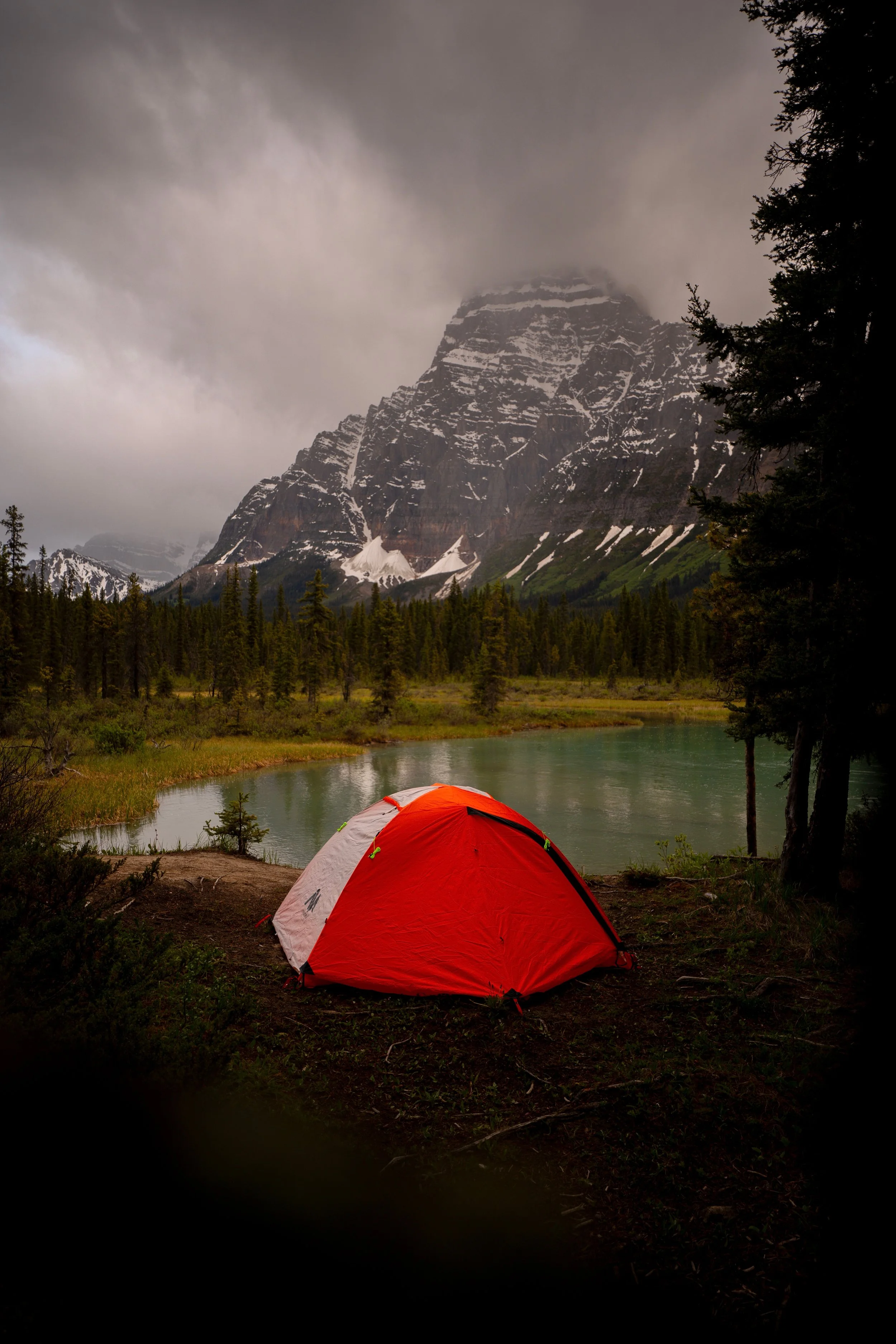 A red and white camping tent set up beside a lake in a forested area with a large mountain in the background, partly covered by clouds.