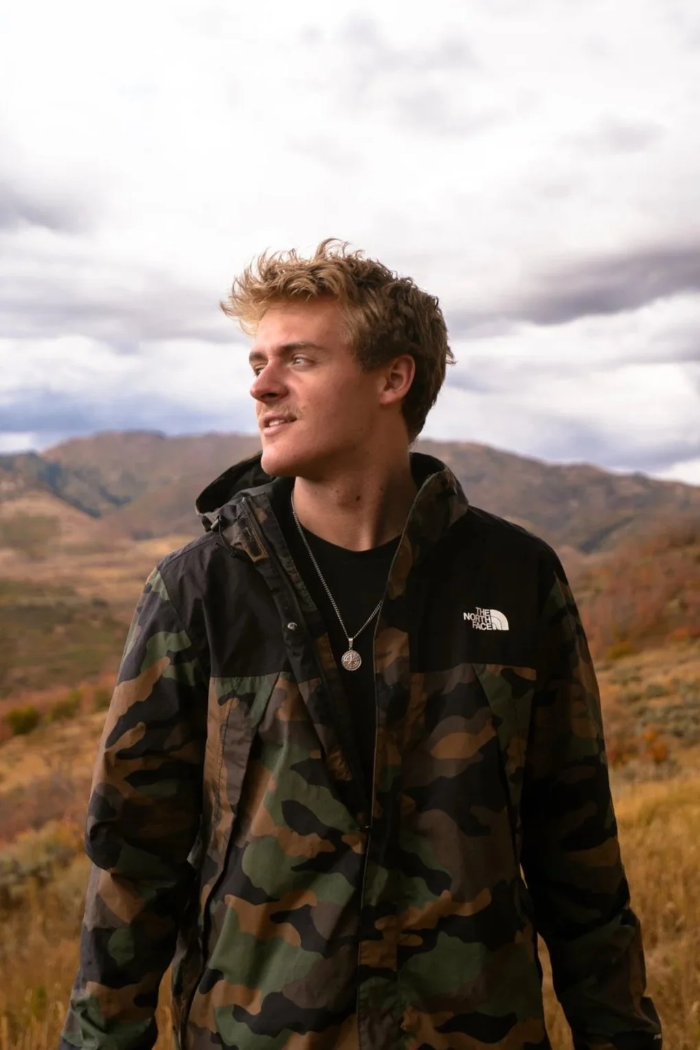 A young man with light brown, curly hair is outdoors, wearing a camouflage jacket with The North Face logo and a necklace with a circular pendant, looking to his left at the mountainous landscape and cloudy sky.