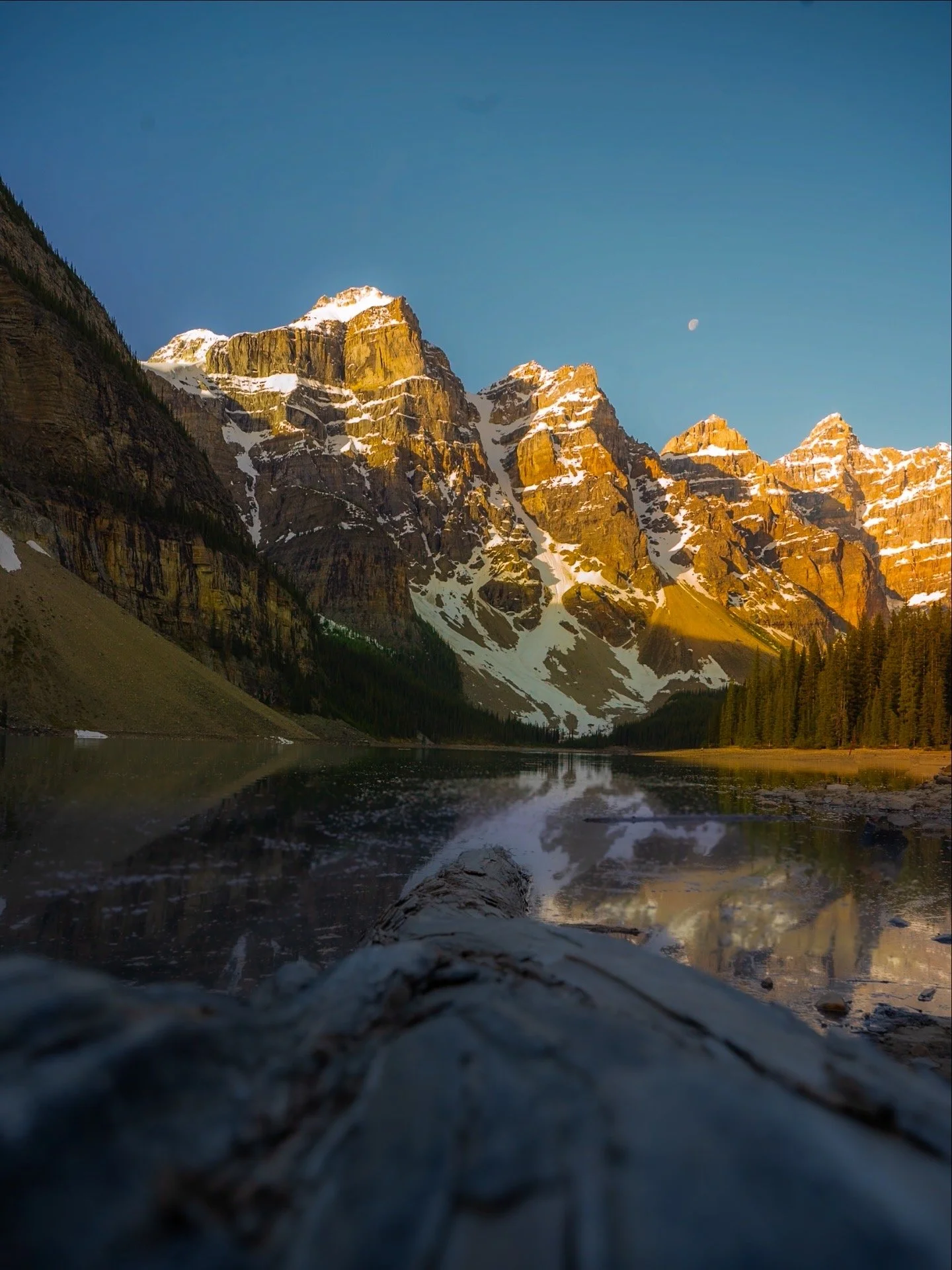 Scenic mountain view with snow-capped peaks reflecting in a lake, trees along the shoreline, and a clear blue sky with the moon visible.