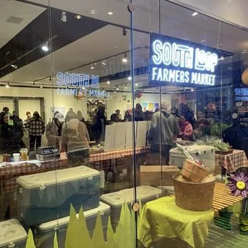 Interior view of a farmers market with shoppers, illuminated 'South Lake Farmers Market' sign, and produce tables.