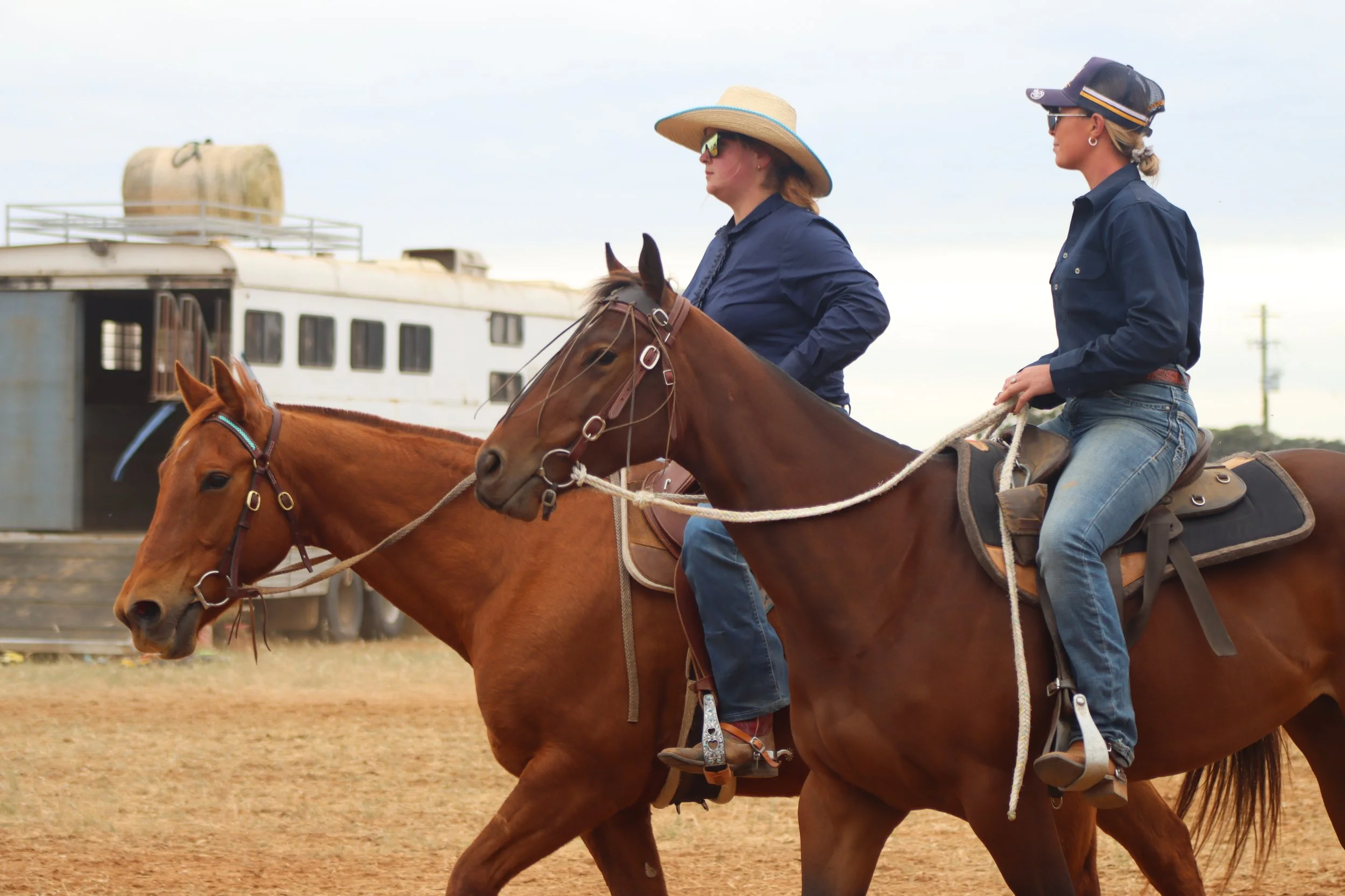 Two women riding horses on a dirt field with an animal trailer and cloudy sky in the background.