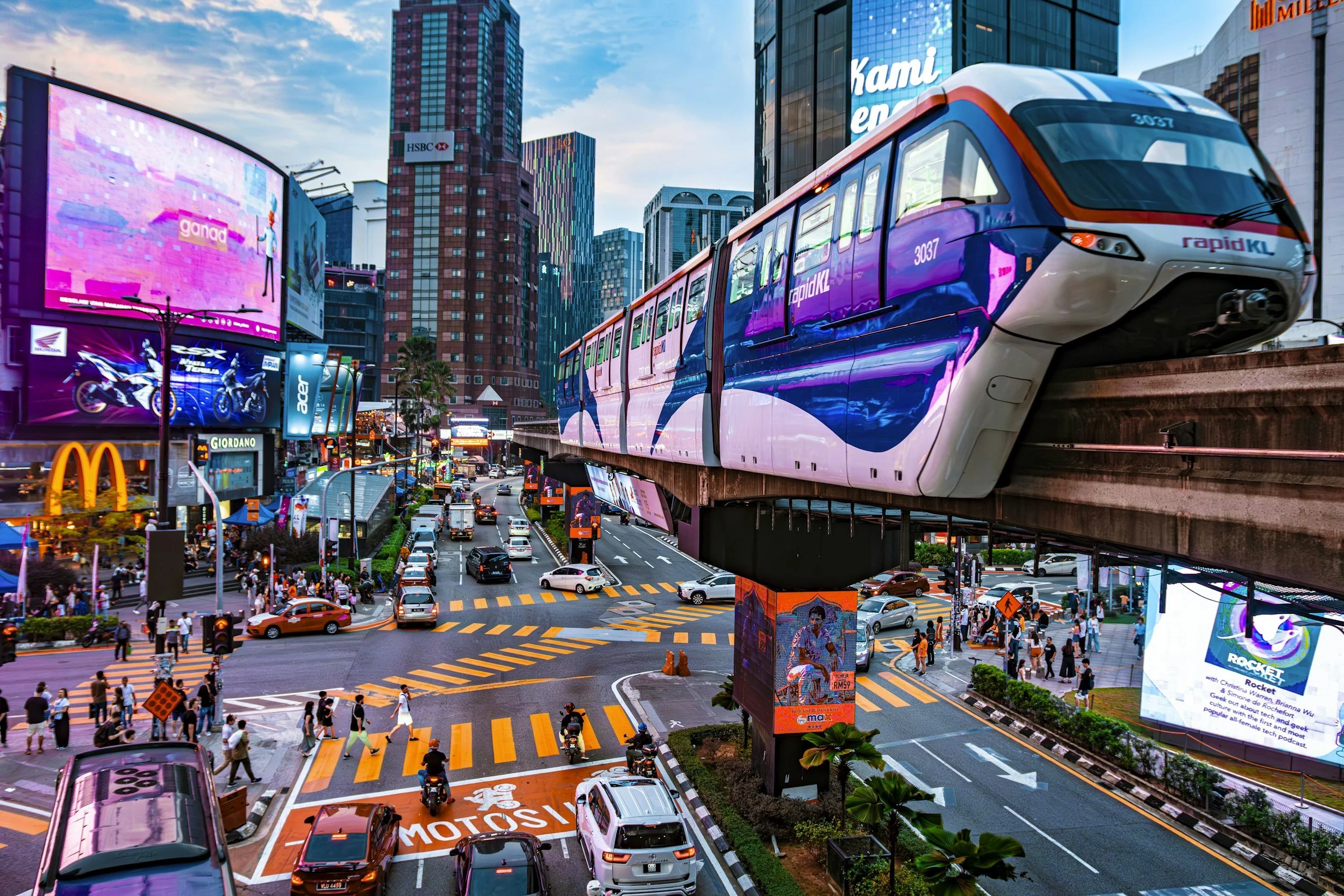 A vibrant city scene with a monorail train passing above busy streets filled with cars, motorcycles, and pedestrians. Tall buildings, billboards, and digital displays adorn the cityscape, with a mix of colorful lights and advertisements, including a McDonald's and various commercial signs.