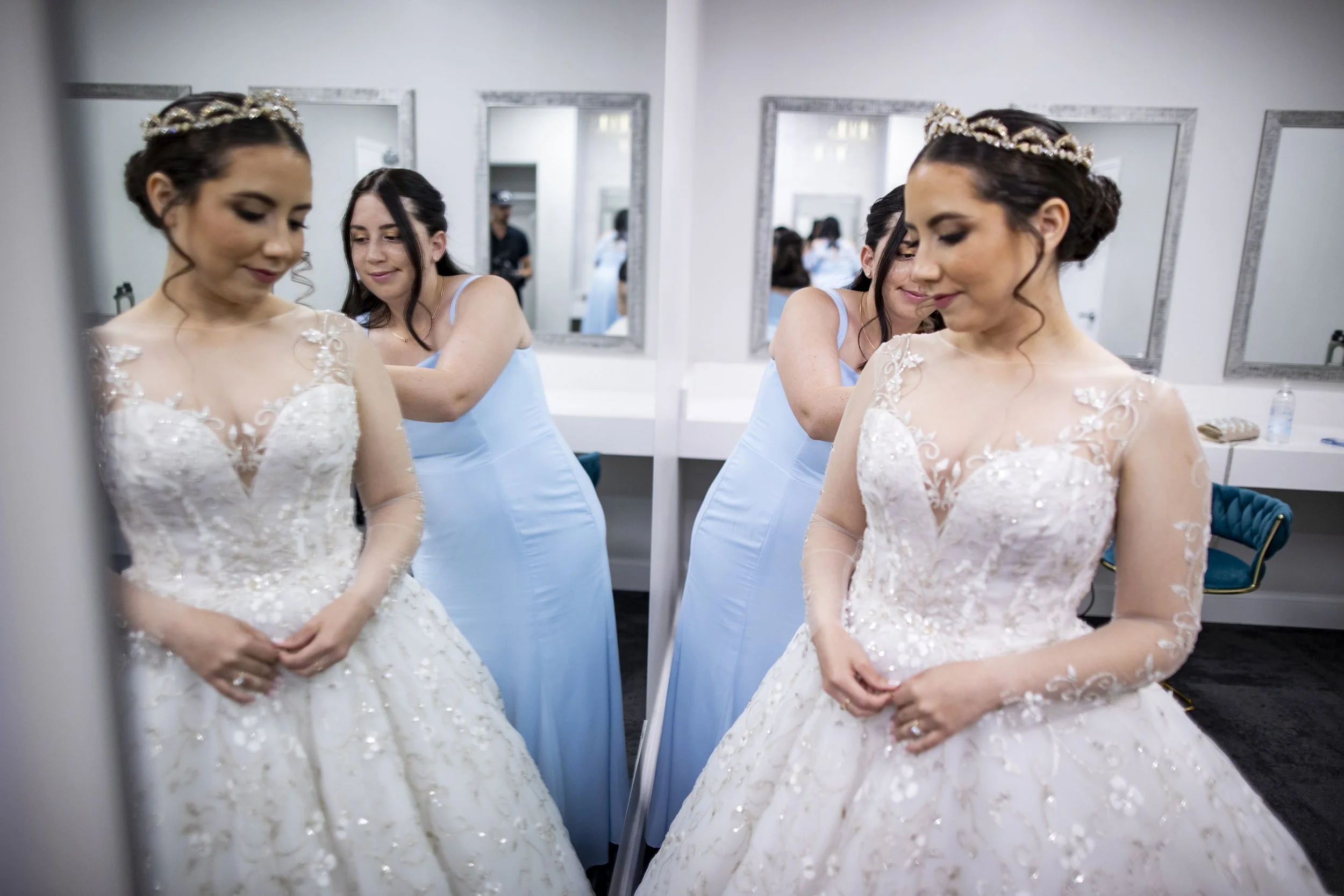 A bride in a white wedding gown with lace and floral details, standing in front of a mirror, getting ready with her bridesmaid in a blue dress adjusting her dress.
