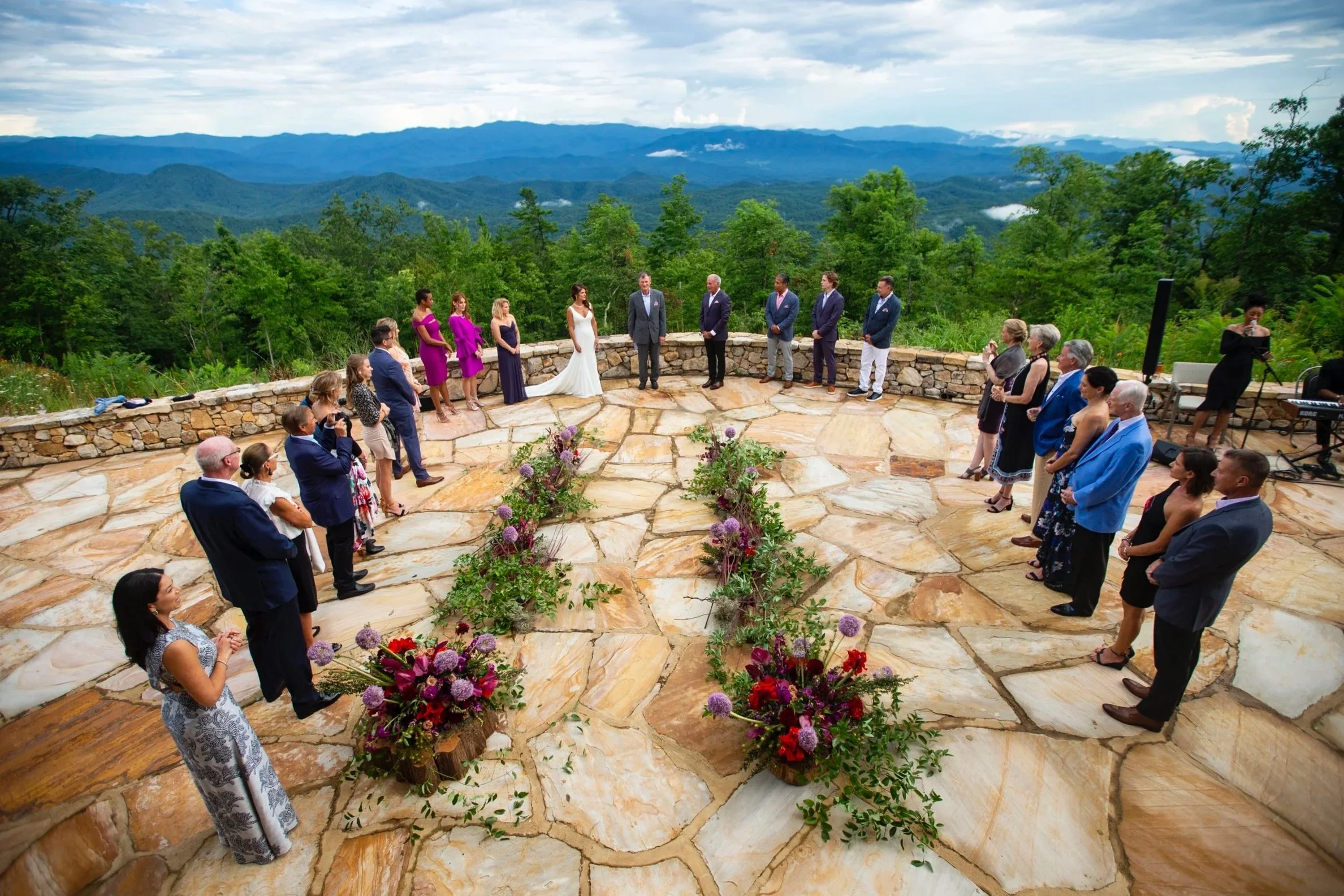 A wedding ceremony taking place on a stone terrace with a scenic mountain view in the background. The bride and groom are standing at the center, surrounded by family and friends.