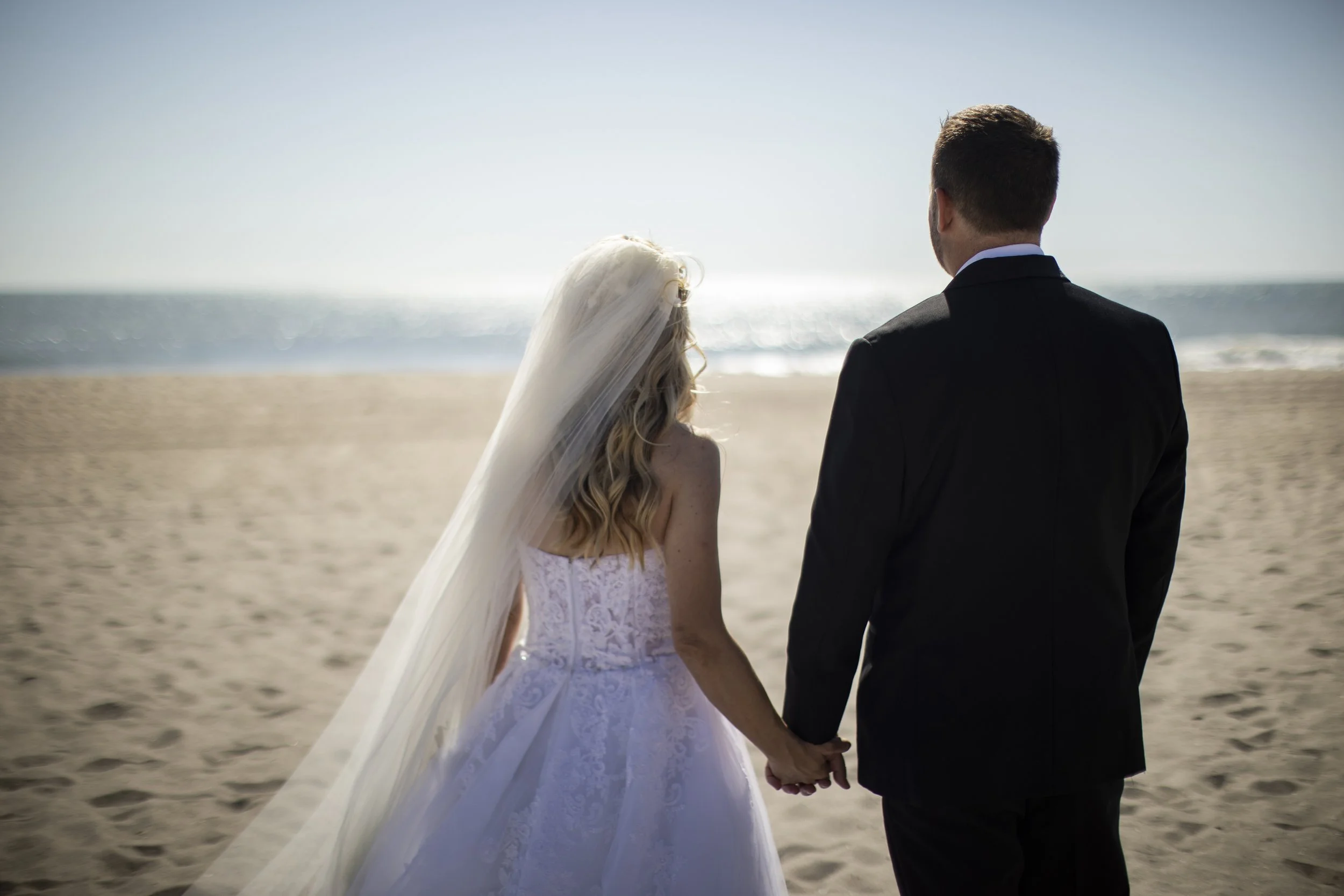 A bride and groom holding hands on a beach, facing the ocean, during their wedding.