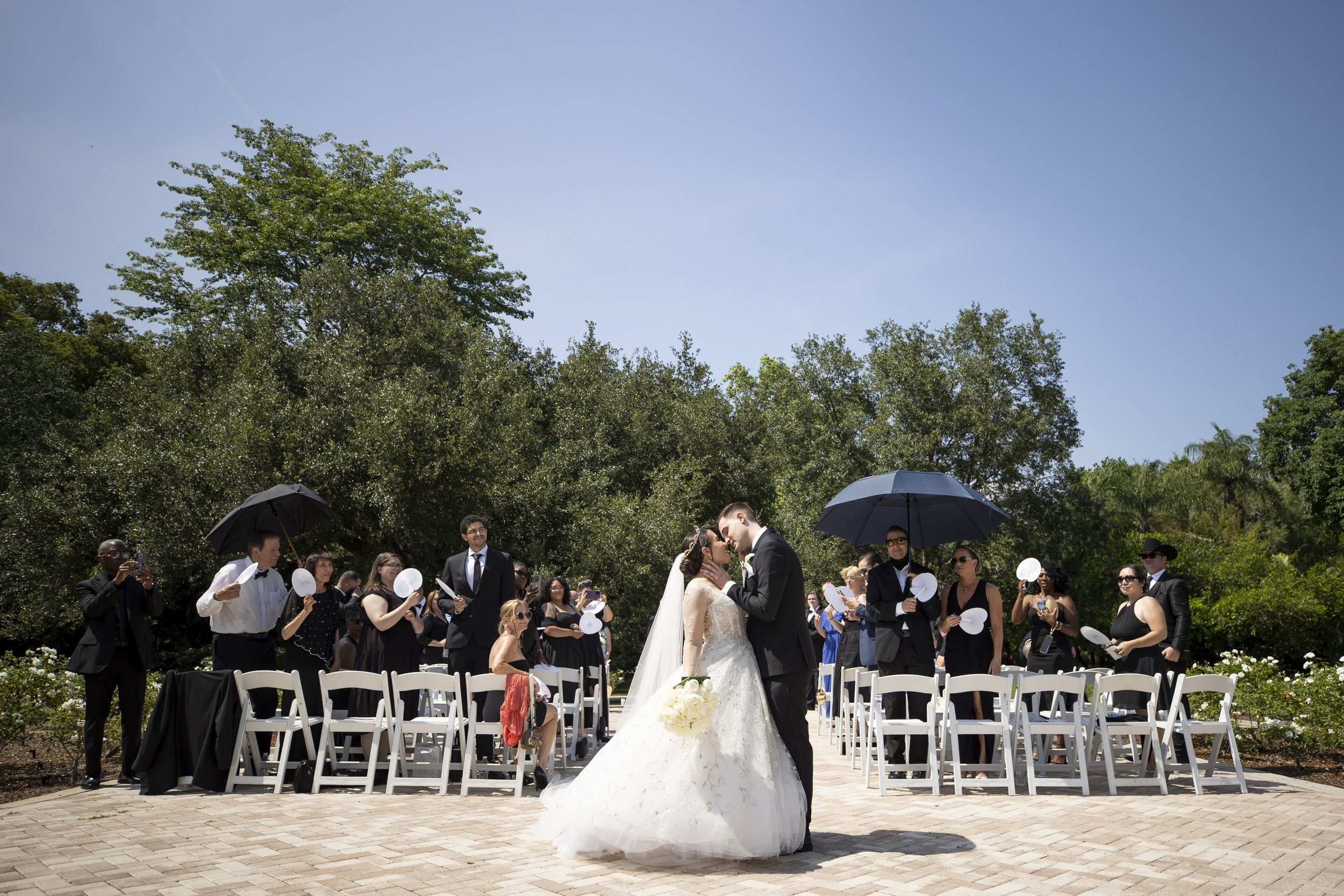 A bride and groom kiss during their outdoor wedding ceremony, surrounded by guests seated on white chairs, some holding umbrellas to shield from the sun, with green trees and a clear blue sky in the background.