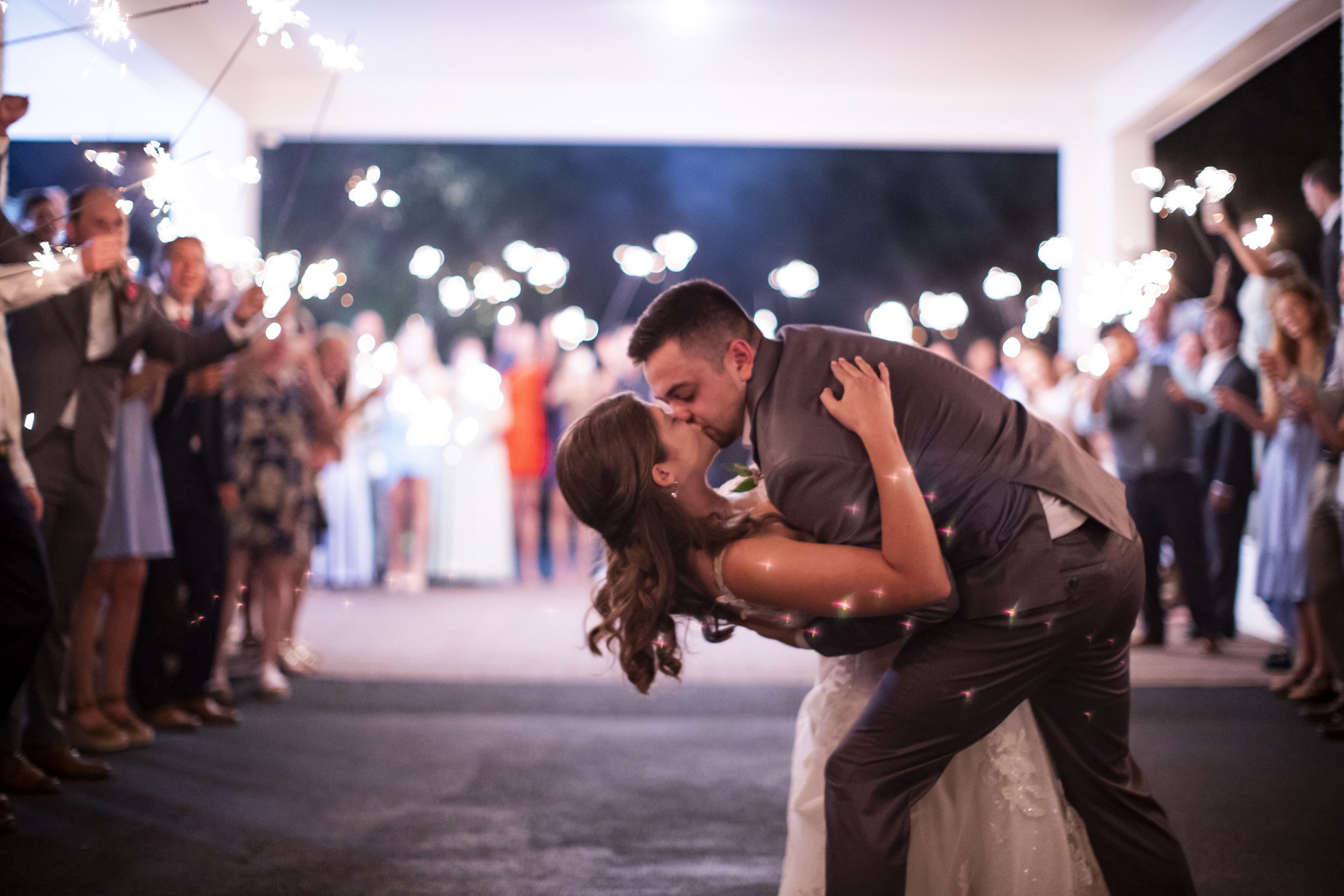 A newlywed couple dancing at their wedding reception, surrounded by guests holding sparklers at night.