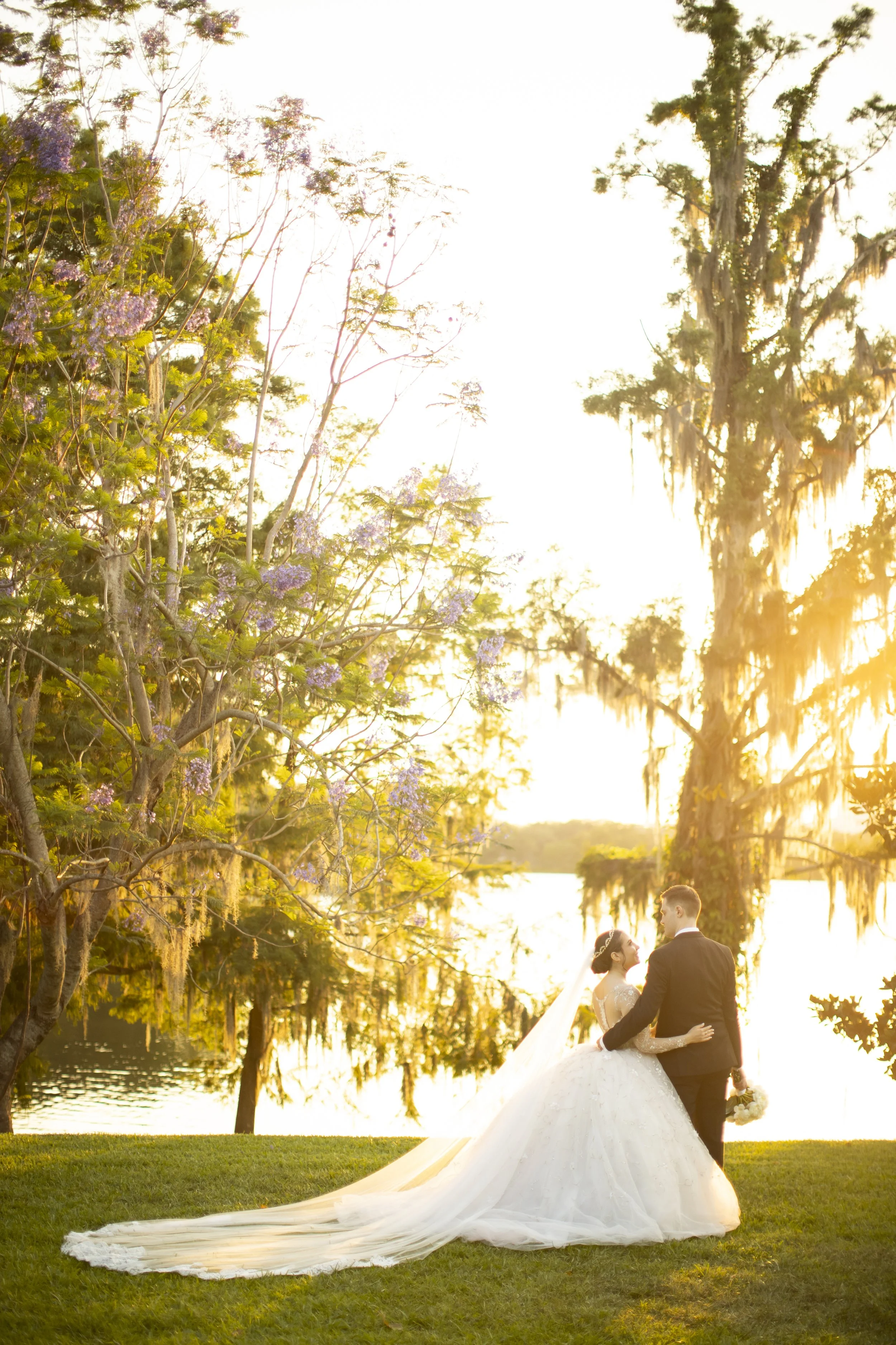 A bride and groom standing on grass near a river during sunset, surrounded by trees, with the bride in a white wedding gown and a long train, and the groom in a black tuxedo.