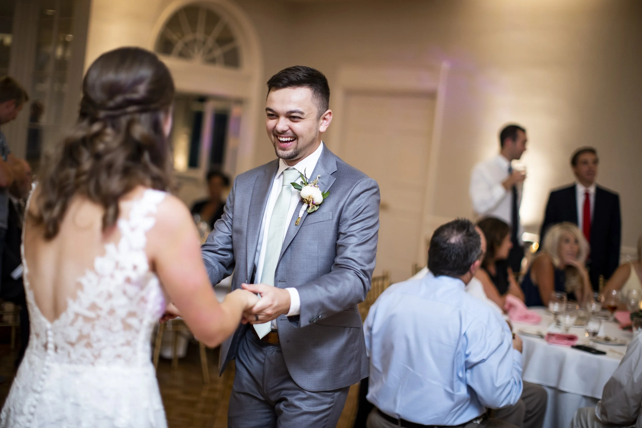 A man in a gray suit and a woman in a white lace dress holding hands and smiling at each other during a wedding reception.