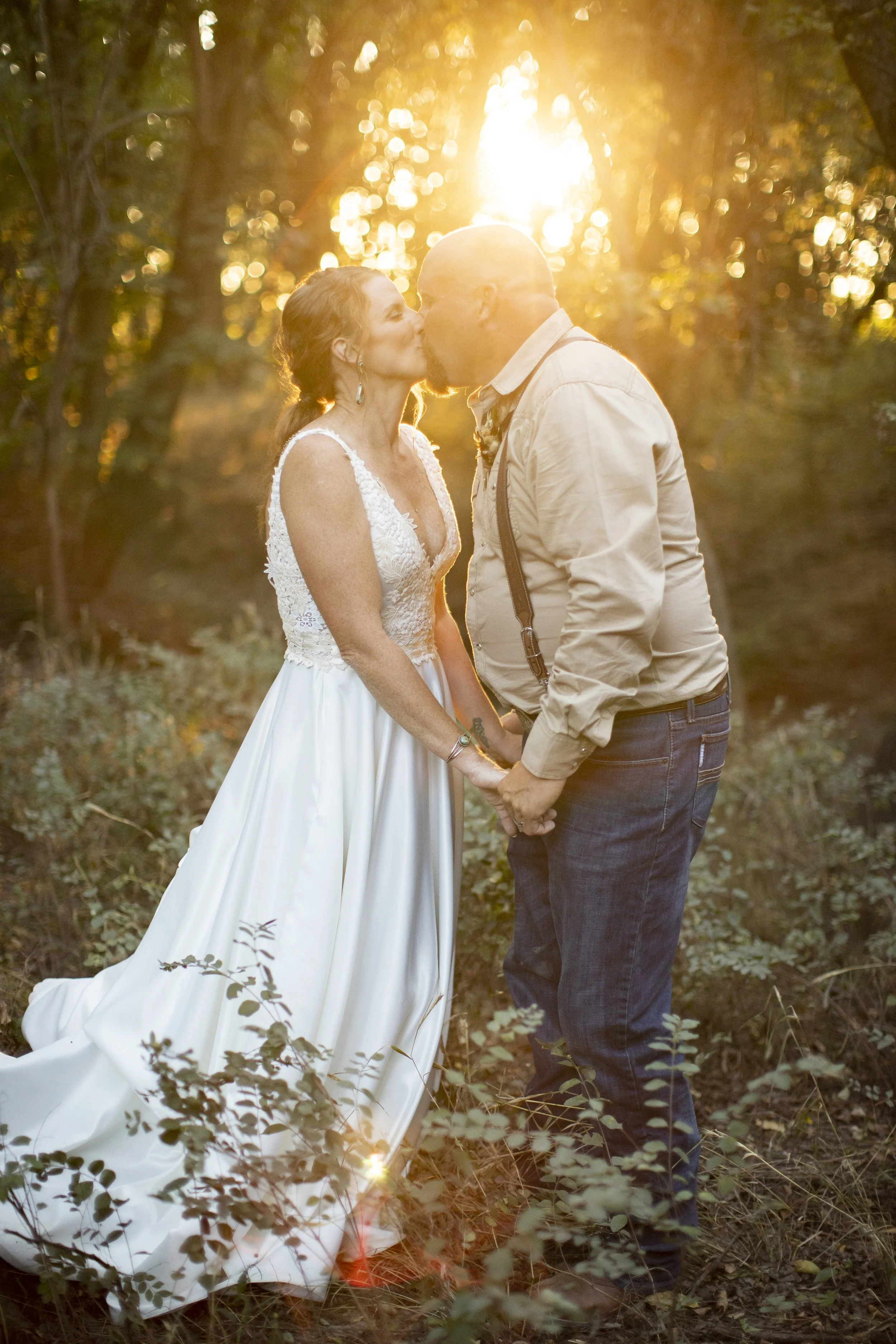 A couple kissing in a forest during sunset, woman in a white wedding dress and man in a casual shirt and suspenders.