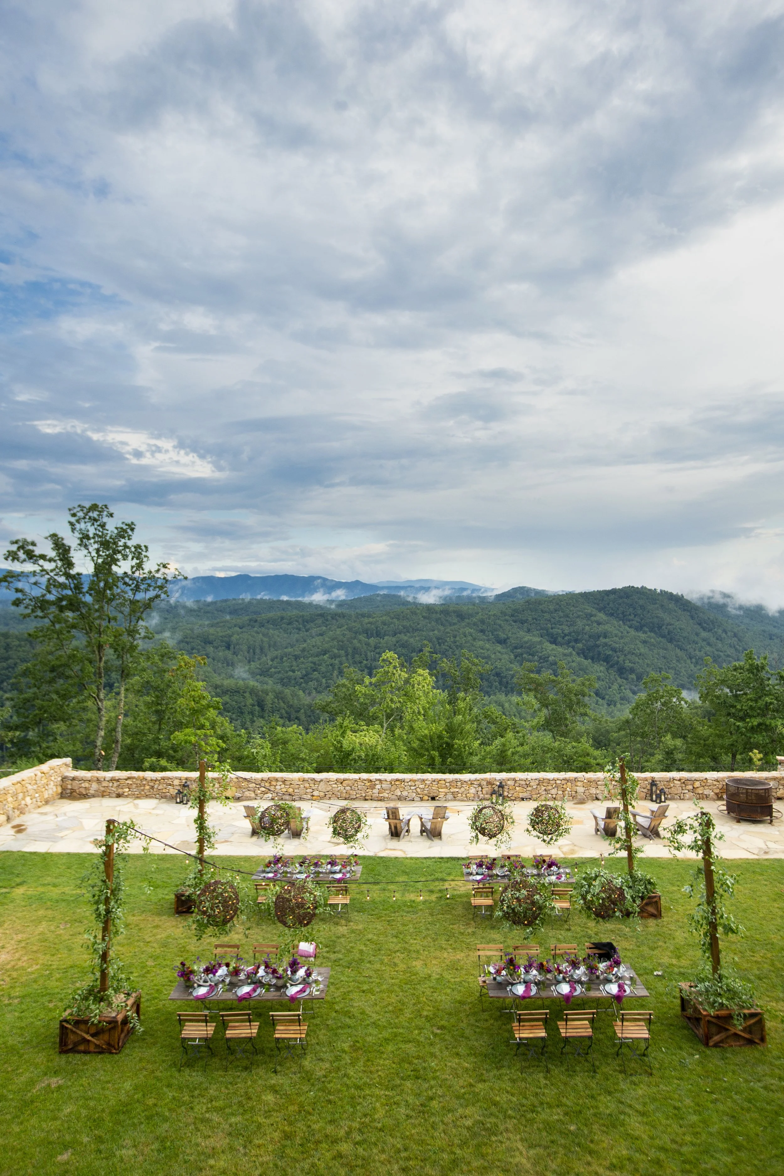Decorative outdoor event setup with tables, chairs, and floral arrangements against a backdrop of green hills and a cloudy sky.
