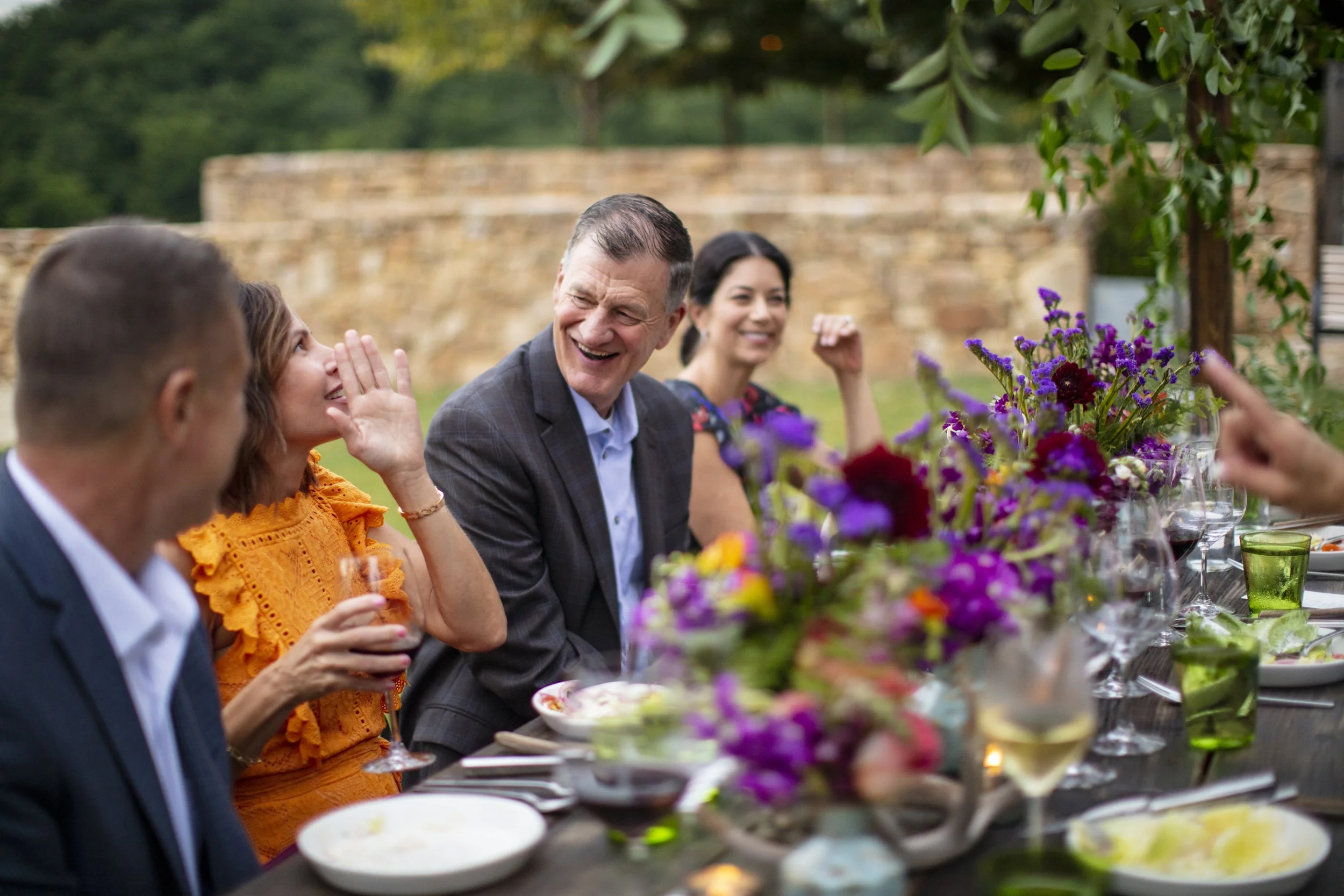 People sitting at a long outdoor dining table, smiling and talking, with a floral centerpiece and glasses of wine, during a garden party or outdoor celebration.