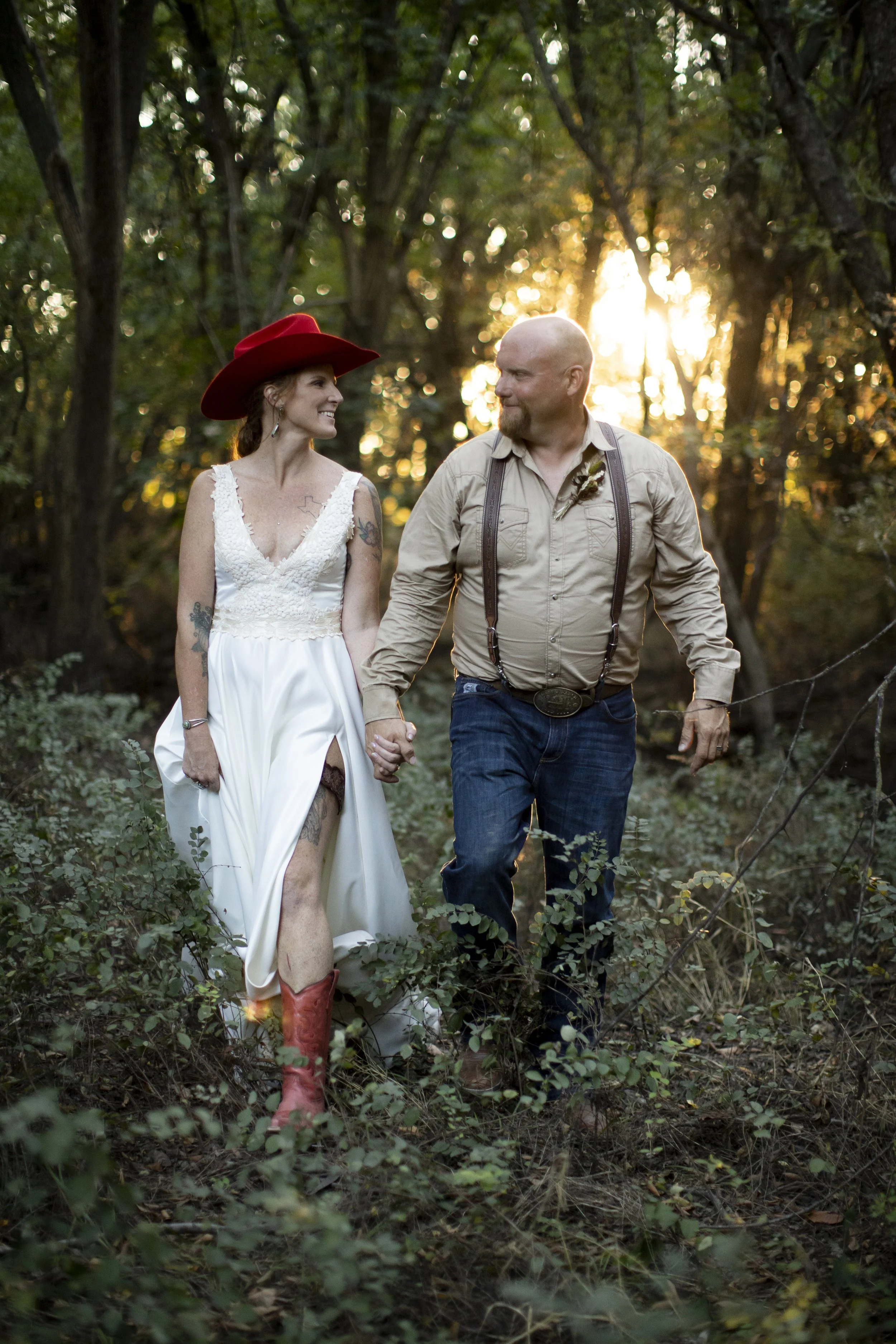 A couple walking hand-in-hand through a wooded area during sunset. The woman is wearing a white dress, cowboy boots, and a large red hat. The man is dressed in casual attire with suspenders. They are smiling at each other.