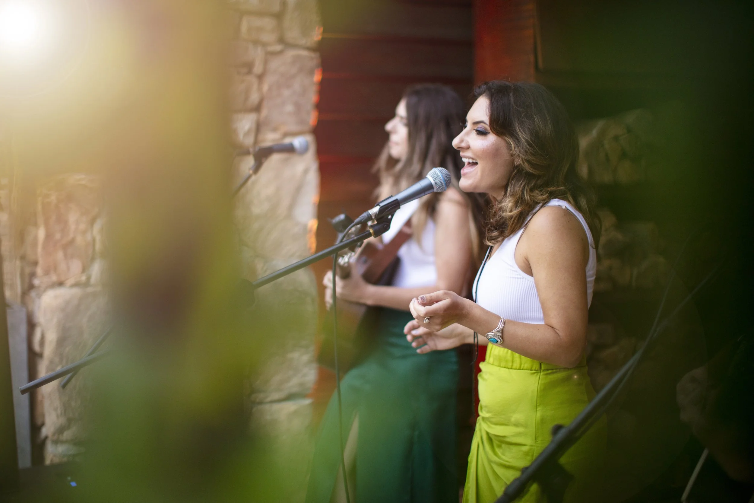 Two women singing into microphones in a rustic indoor setting with a brick wall background, blurred green plants in foreground, one in a white top and lime-green skirt, the other in a white top and dark green skirt.