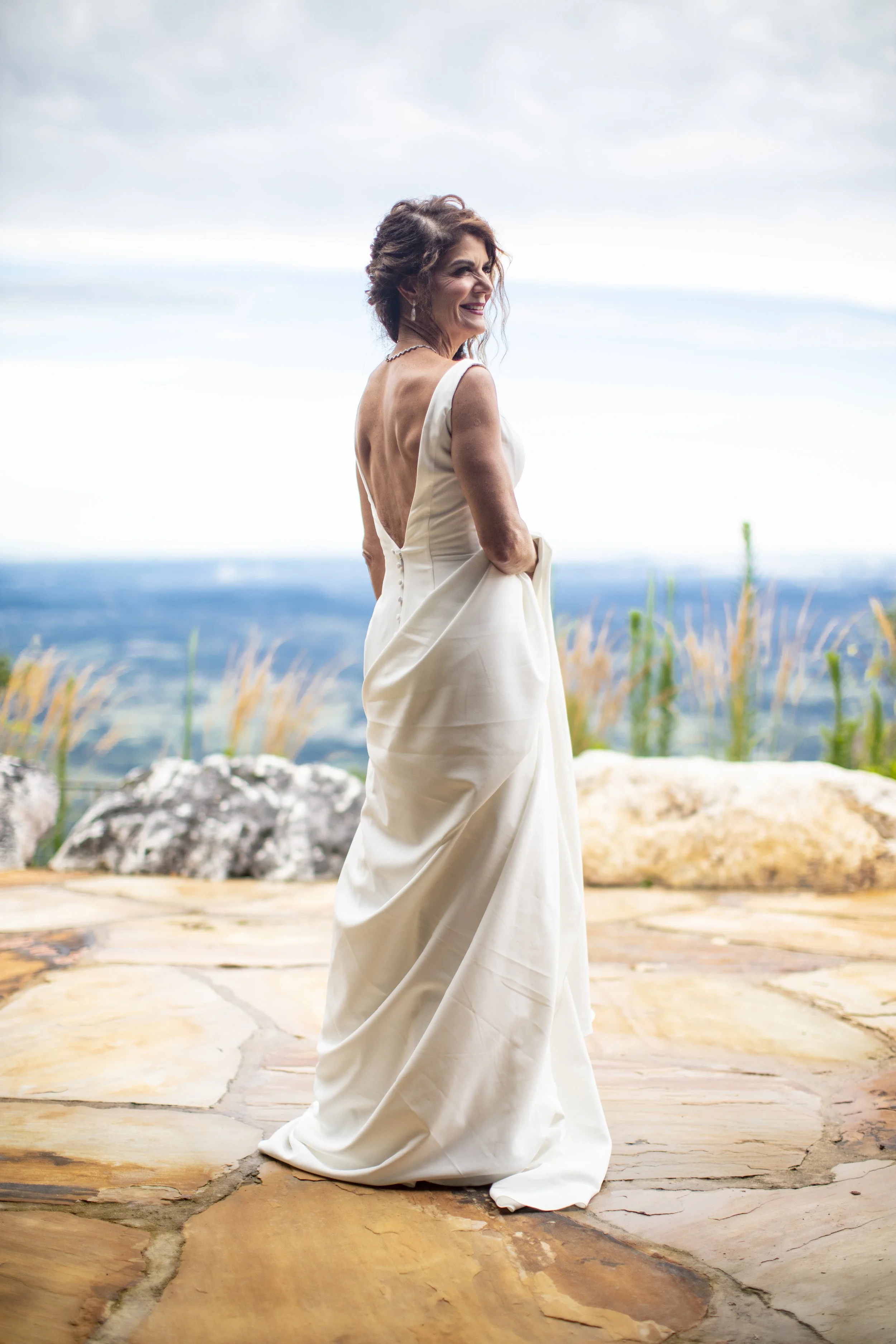 A woman in a white wedding dress standing outdoors on a stone patio, smiling and looking away, with a view of a blue sky and distant landscape in the background.