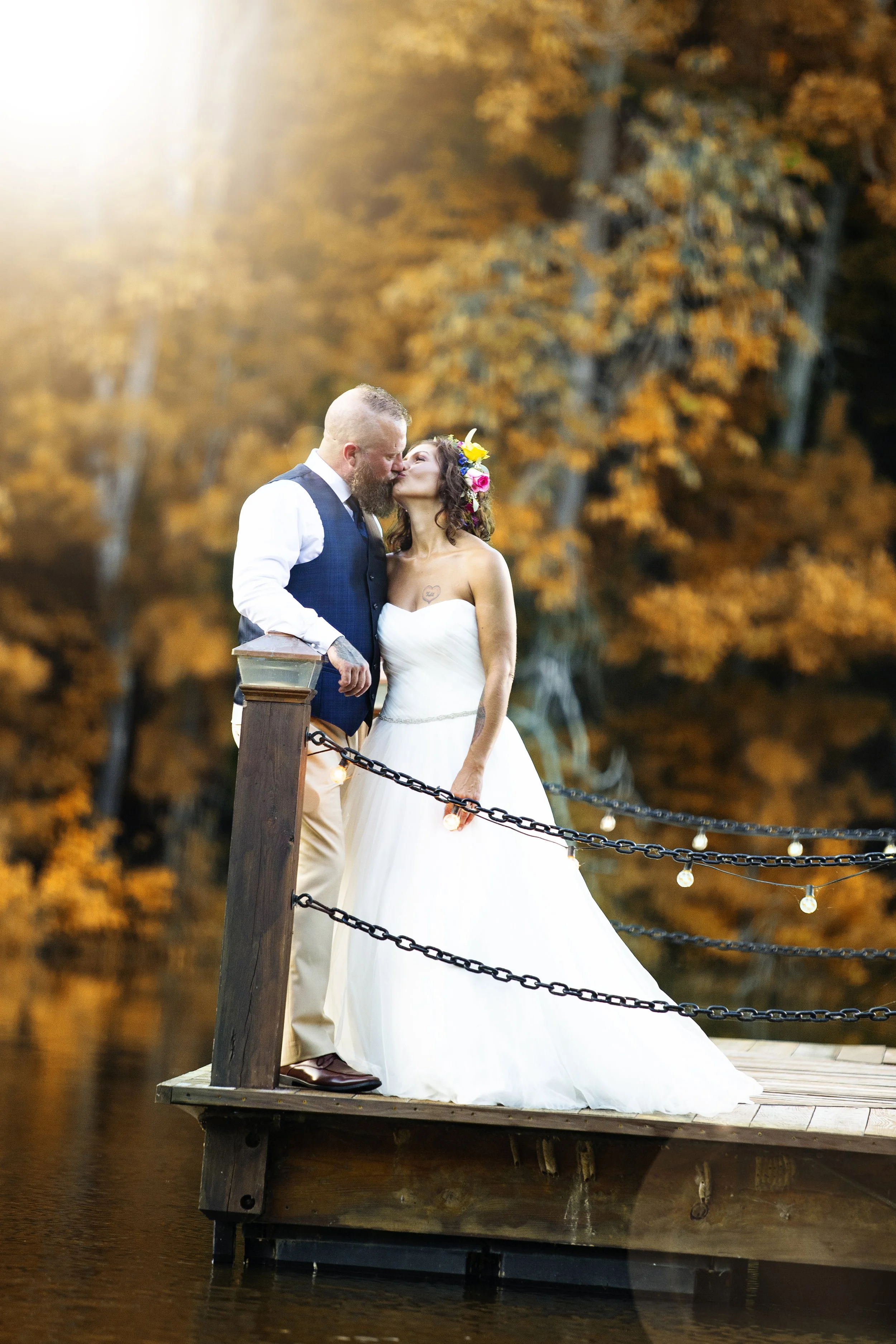 A bride and groom share a kiss on a wooden dock by a lake surrounded by fall foliage, with string lights hanging along the railing, during sunset.