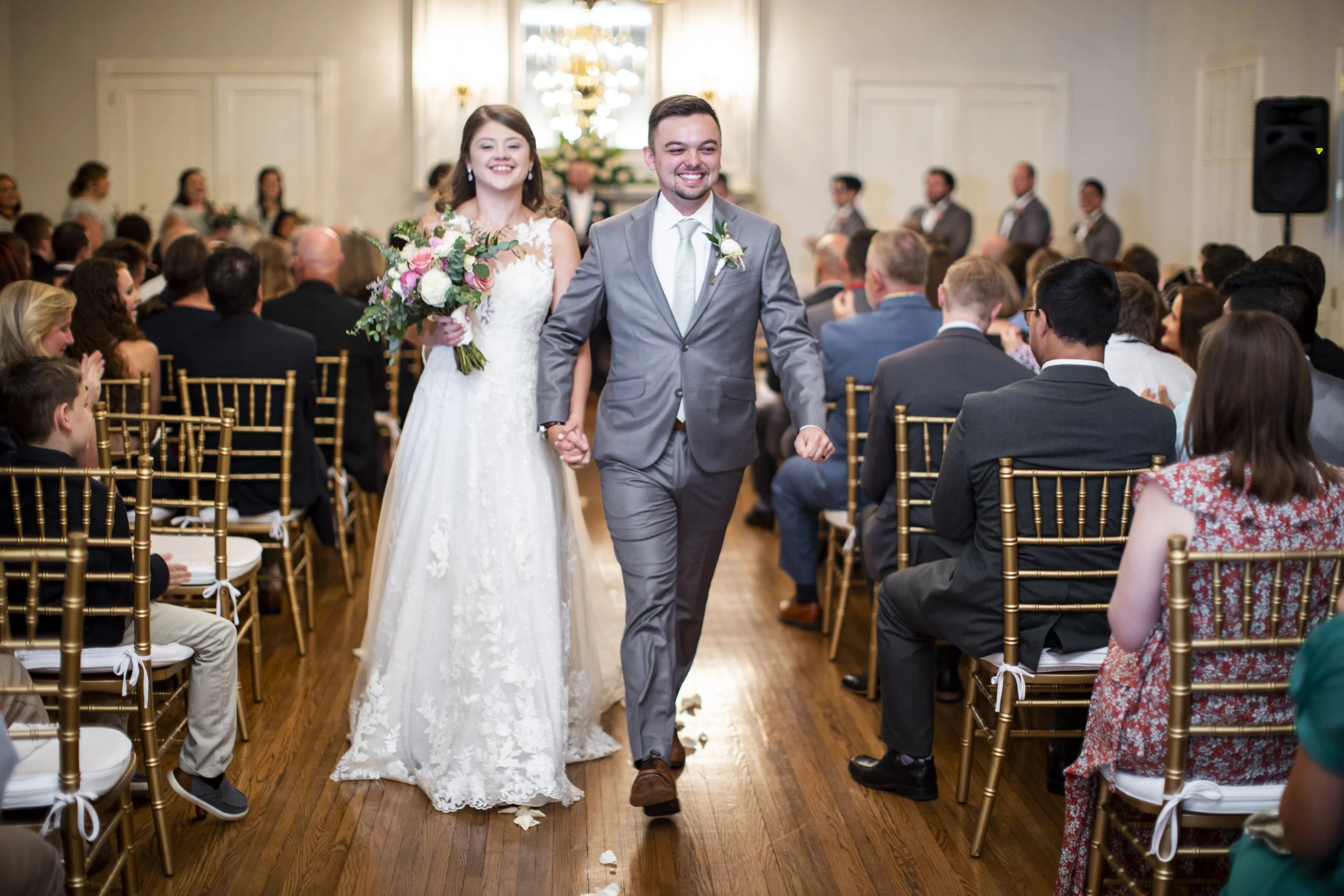 A newlywed couple walking down the aisle holding hands in a wedding ceremony. The bride is wearing a white wedding dress and holding a bouquet, while the groom is in a gray suit. Guests are seated on both sides of the aisle, clapping and smiling.