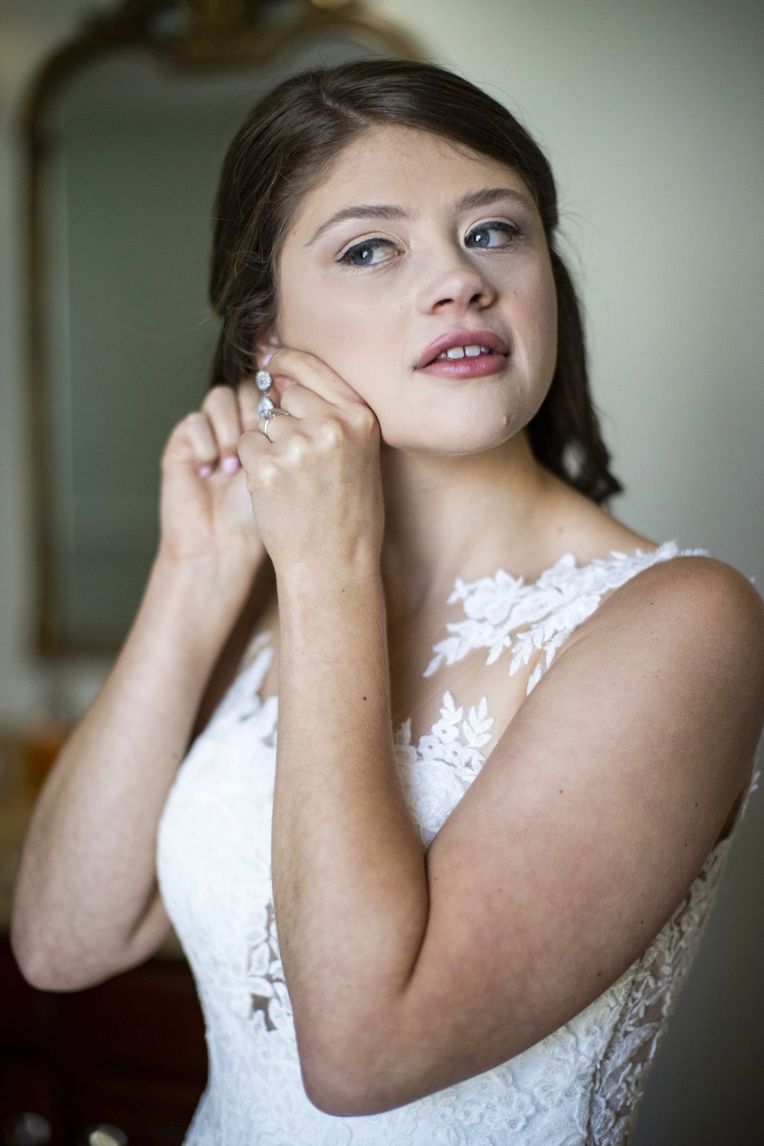 A bride adjusting her earring while looking to the side, wearing a white lace wedding dress, standing in a room with a mirror in the background.