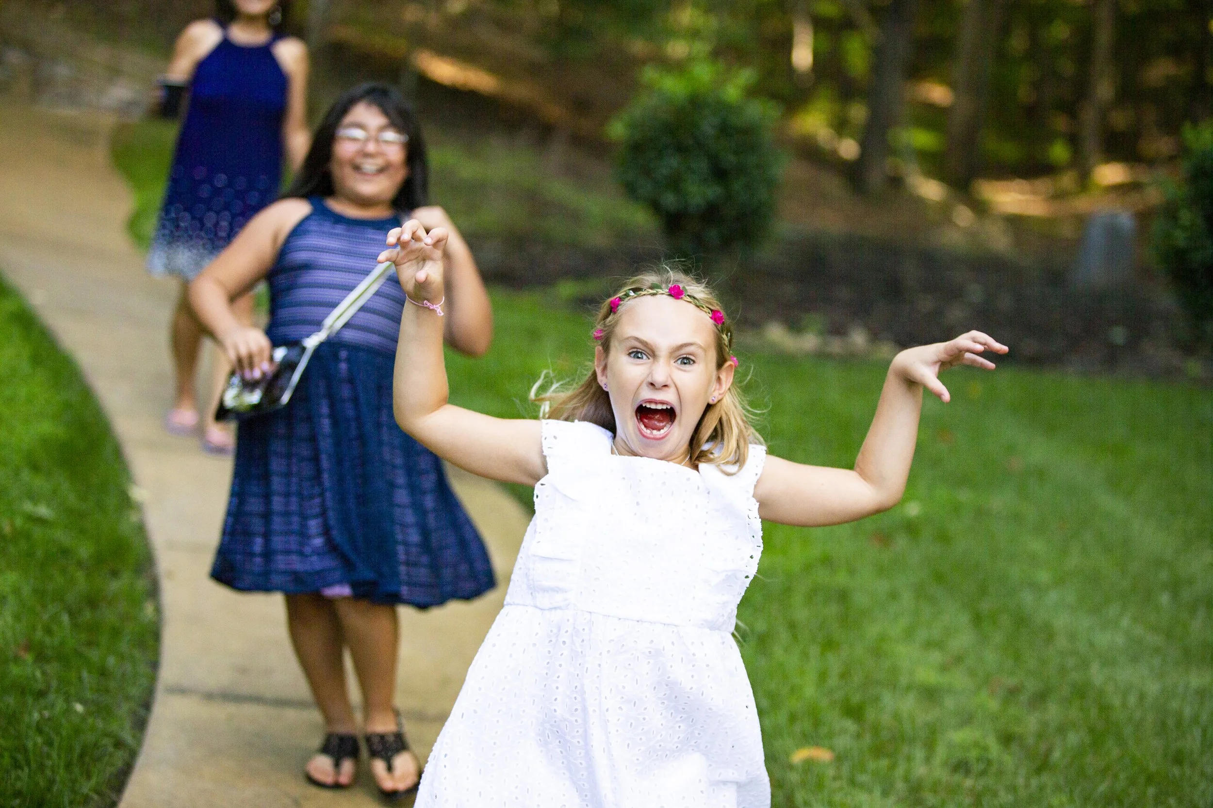Young girl in a white dress with a pink flower headband appears angry and fierce, raising her arms and yelling at the camera, while three other children in blue dresses walk behind her on a grassy pathway in a park.