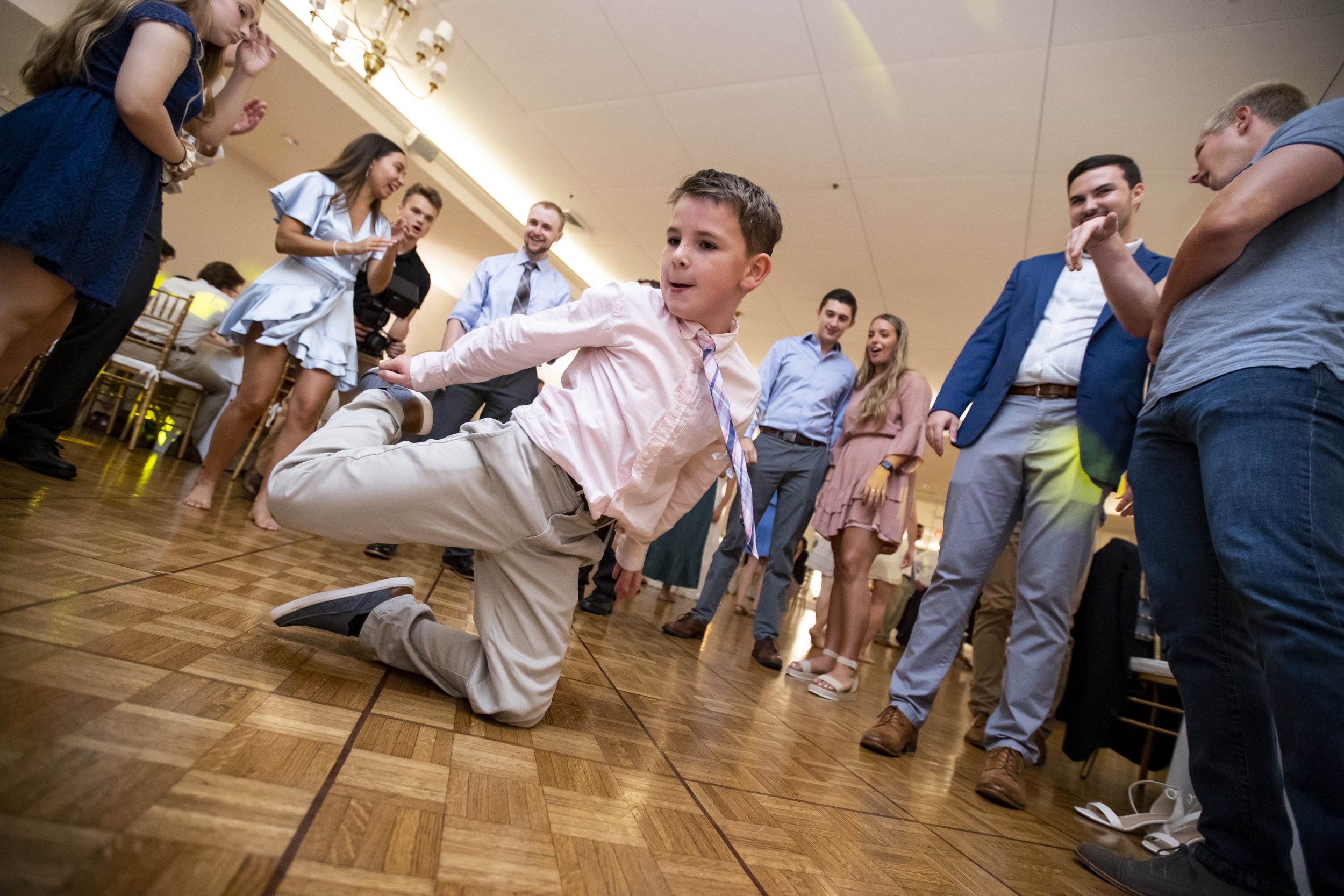 A young boy at a party or dance floor performing a dance move while a group of adults and children watch and smile.