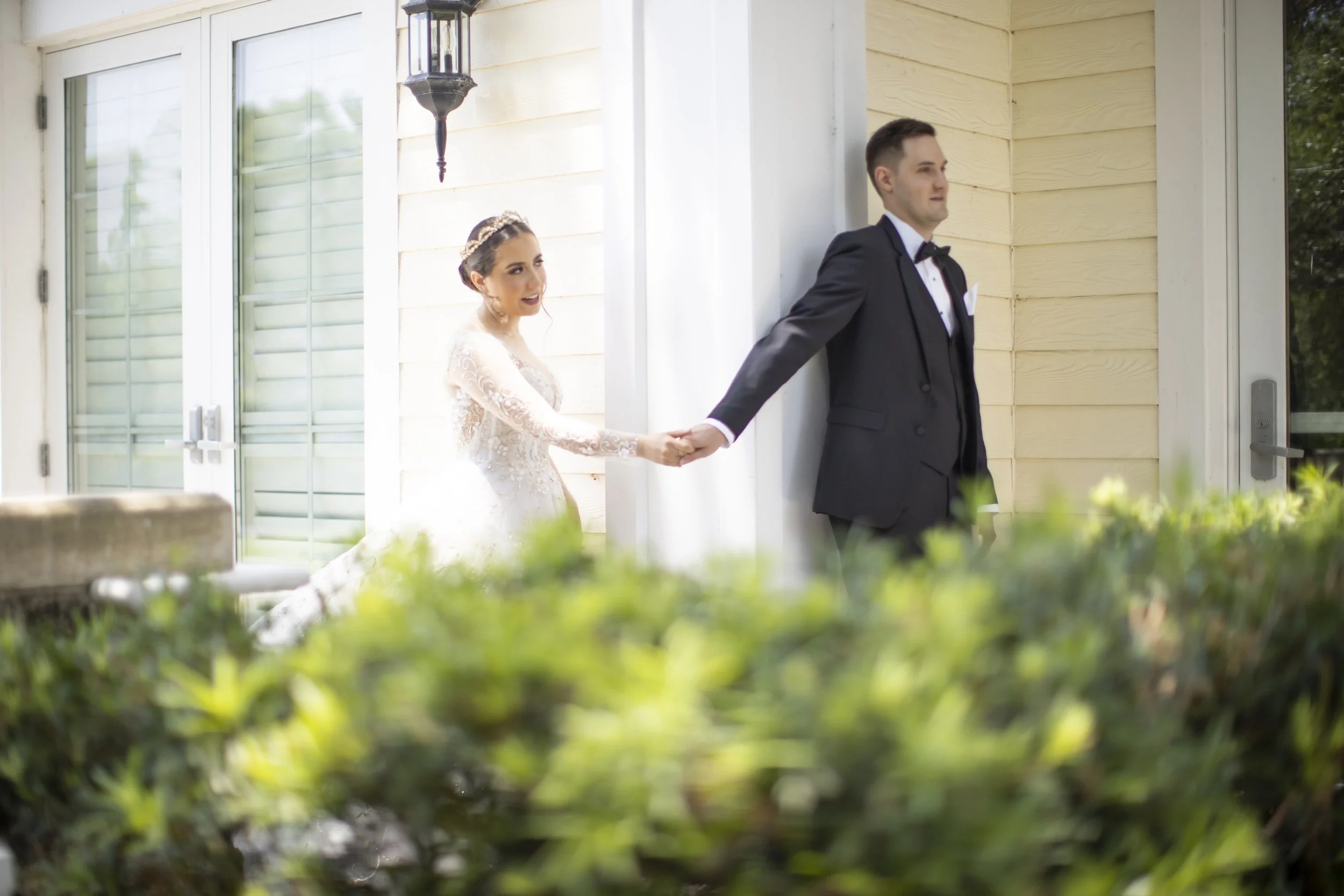A bride in a white lace wedding dress holding hands with a groom in a black tuxedo, standing outside near a house with yellow siding and white trim, with greenery in the foreground.