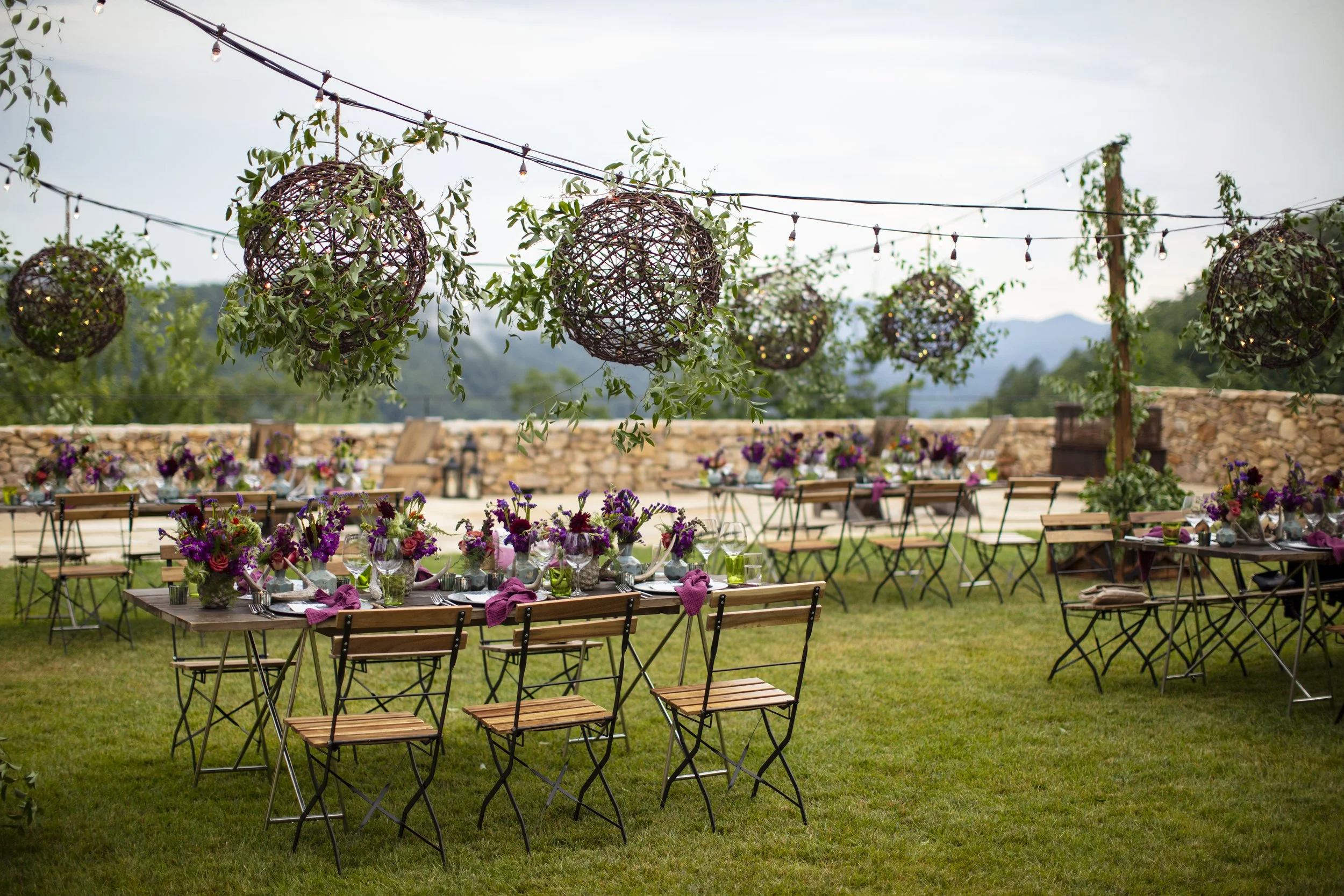 Outdoor event setup with long tables decorated with purple and pink floral centerpieces, purple napkins, and glassware, on a grassy lawn with hanging spherical flower arrangements and string lights overhead.