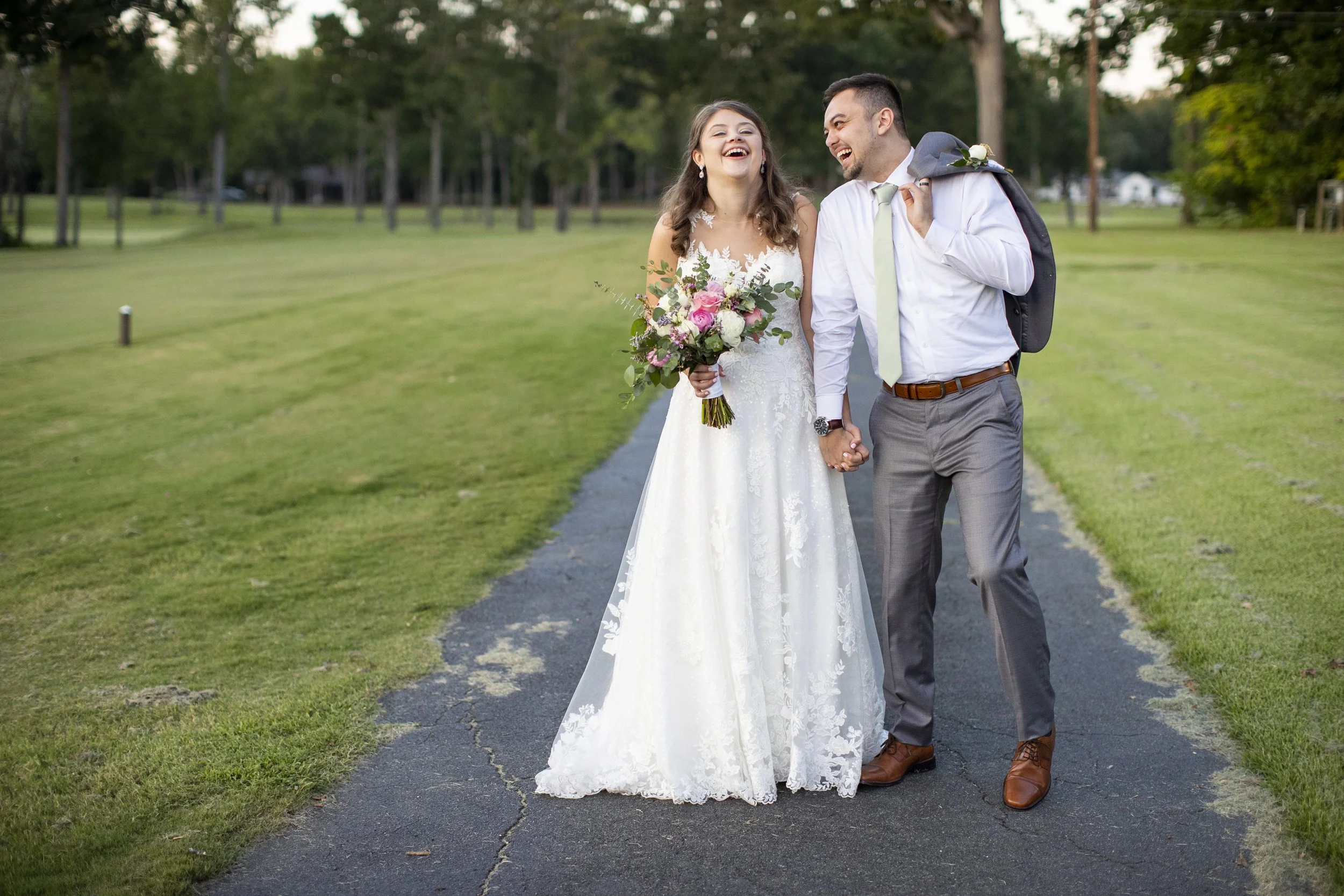 A newlywed couple walking hand in hand on a paved path in a green park, laughing and smiling. The bride wears a white lace wedding dress and holds a bouquet of pink and white flowers. The groom wears a white shirt, tie, gray pants, and carries a suit