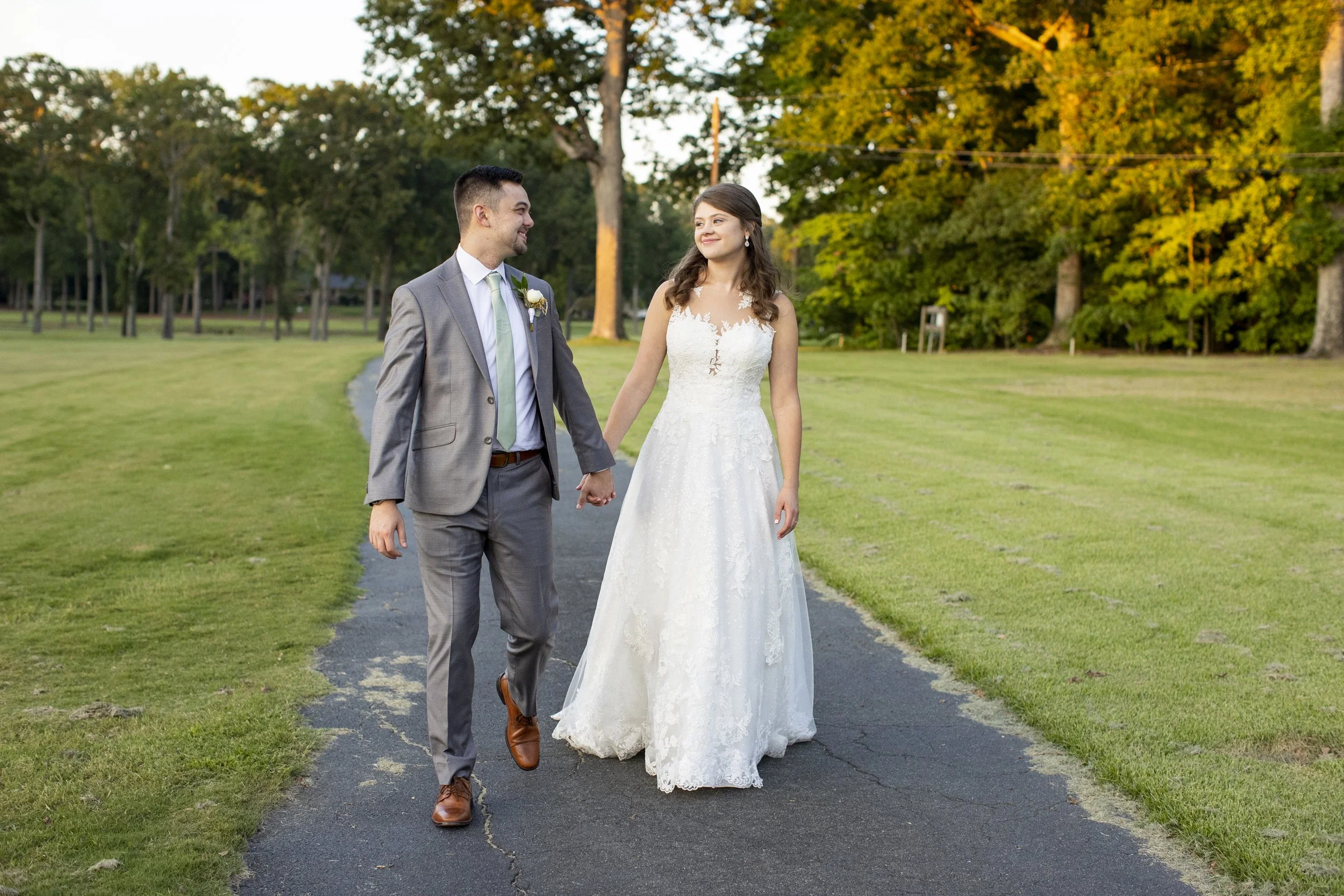 The image shows a bride and groom walking hand-in-hand on a paved path in a park during sunset. The bride wears a white lace wedding dress, and the groom wears a gray suit with a light green tie. They are smiling and looking at each other, with green