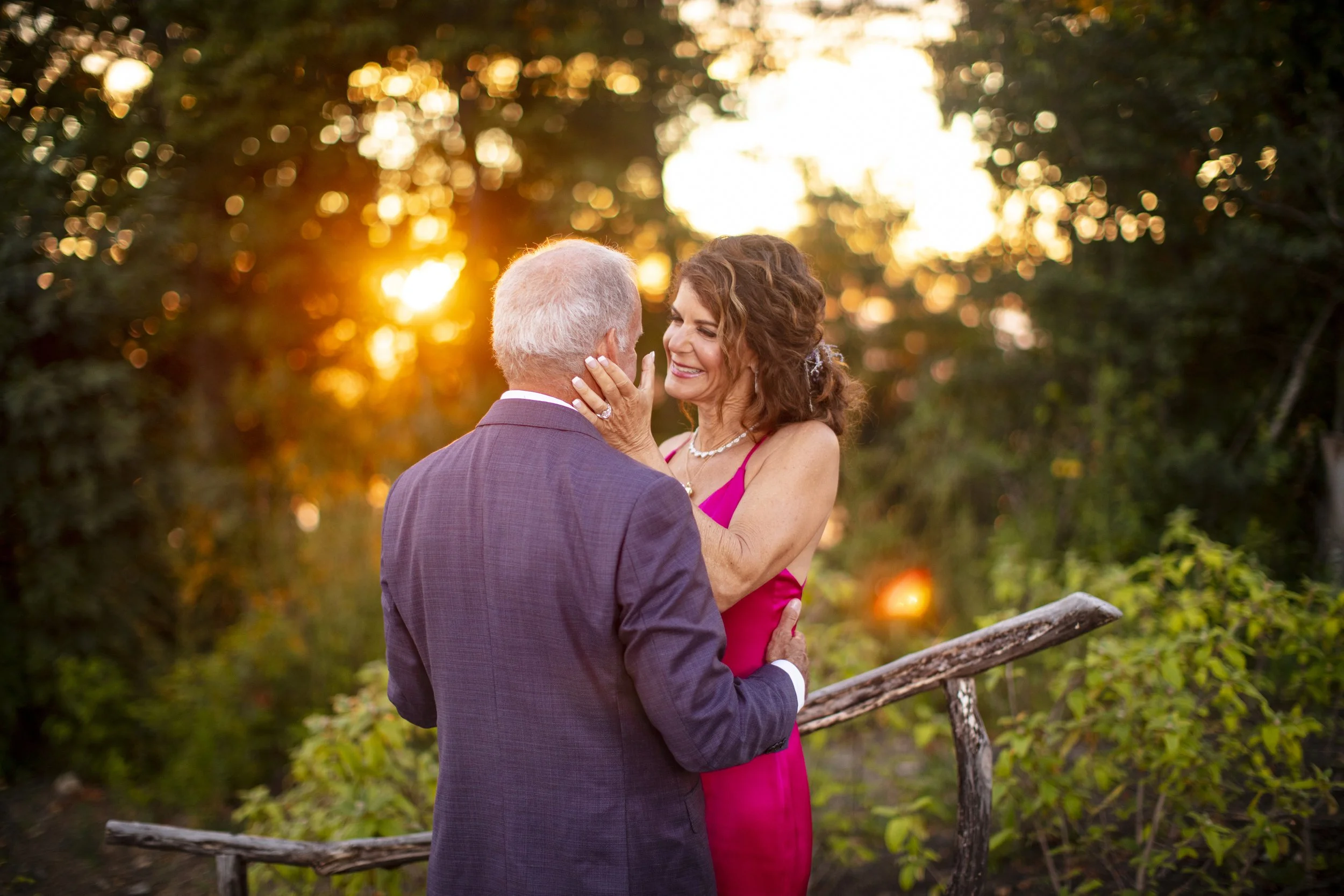 A woman in a pink dress has her hand on an elderly man's face, smiling and gazing at him lovingly during sunset outdoors, with trees and a rustic wooden fence in the background.