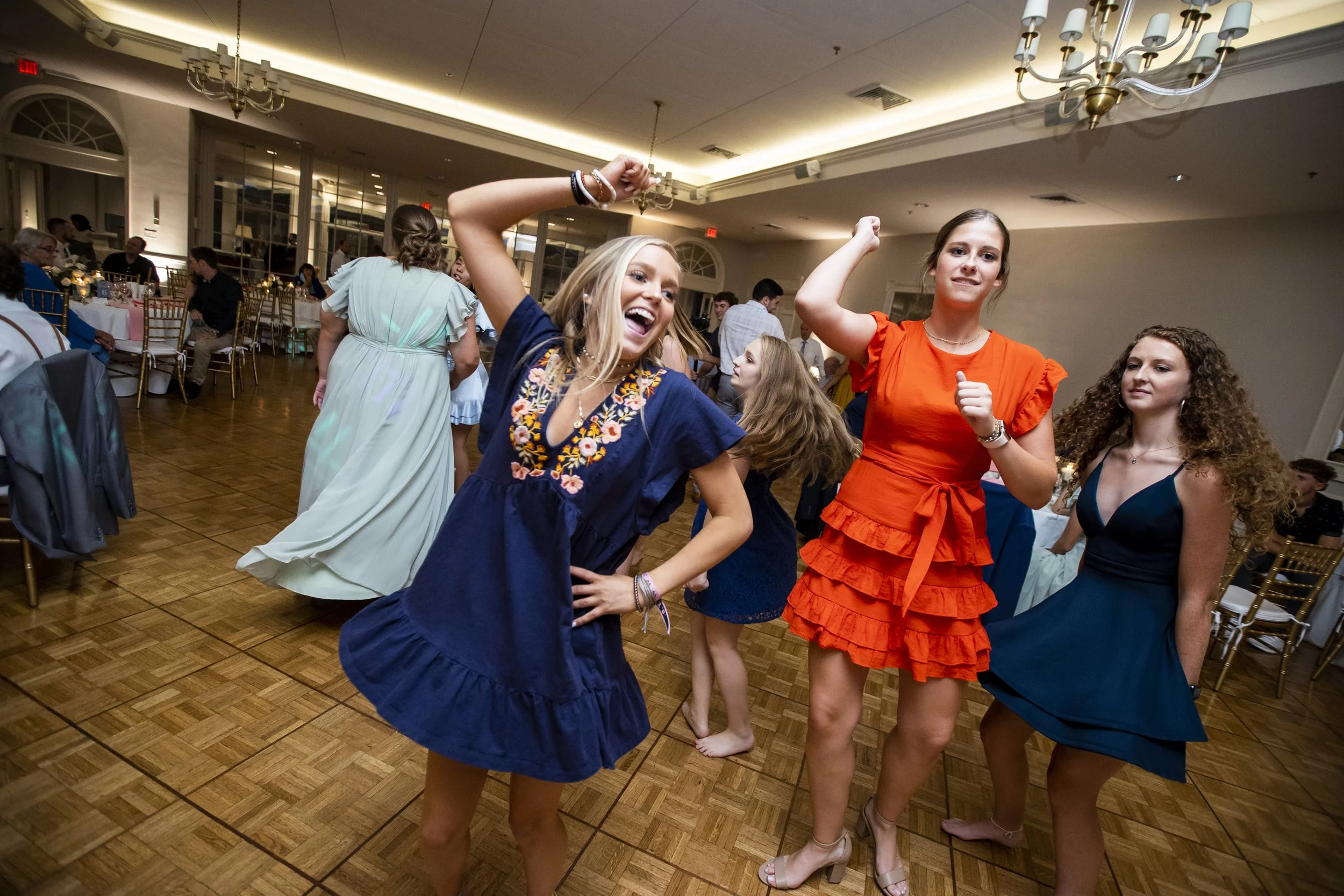 Three women dancing and having fun at a social event in a large decorated banquet hall with tables and guests in the background.