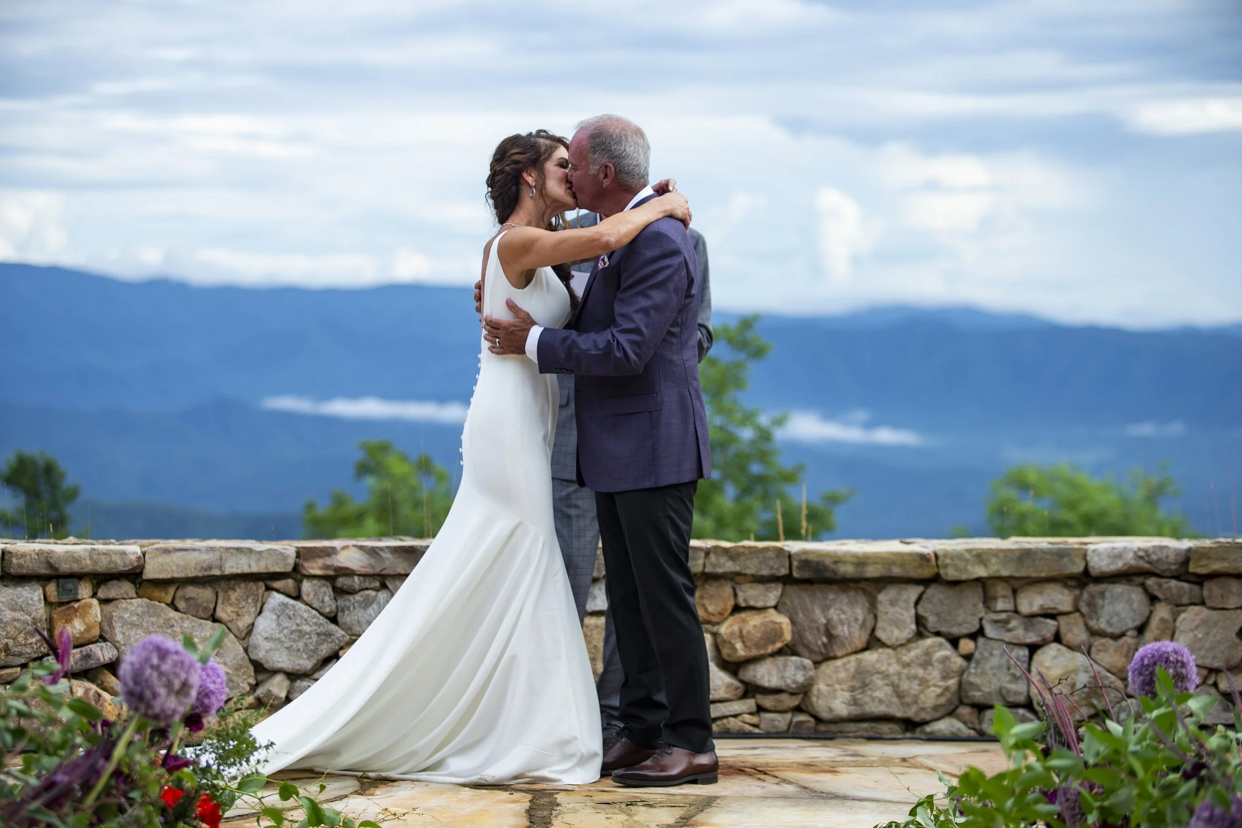 A bride and an older man, possibly her father, sharing a kiss during a wedding ceremony outdoors with mountains and blue sky in the background.
