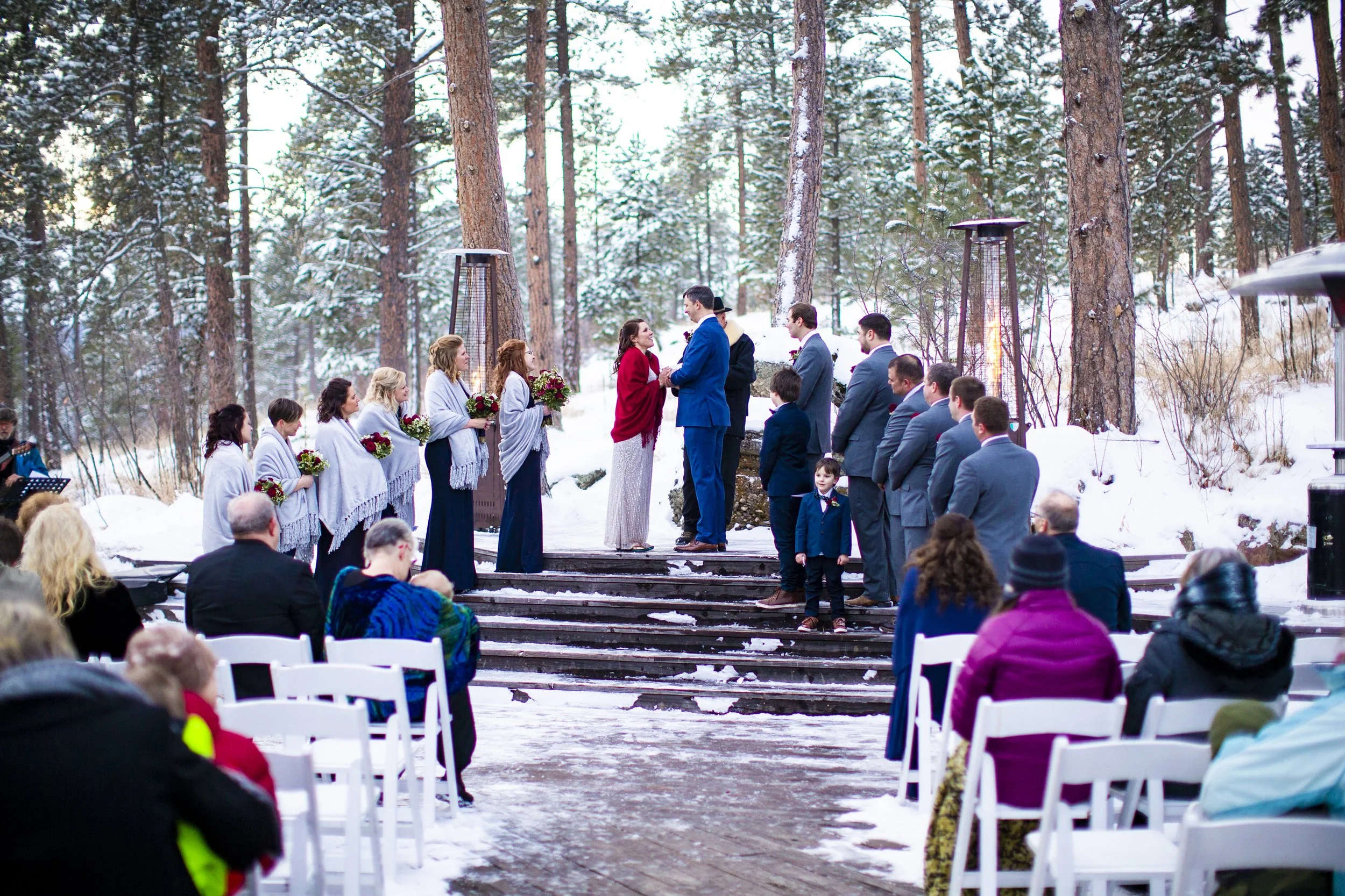 A winter outdoor wedding ceremony with snow-covered ground and tall pine trees in the background. The bride and groom stand at the altar surrounded by bridesmaids, groomsmen, and guests seated on white chairs.